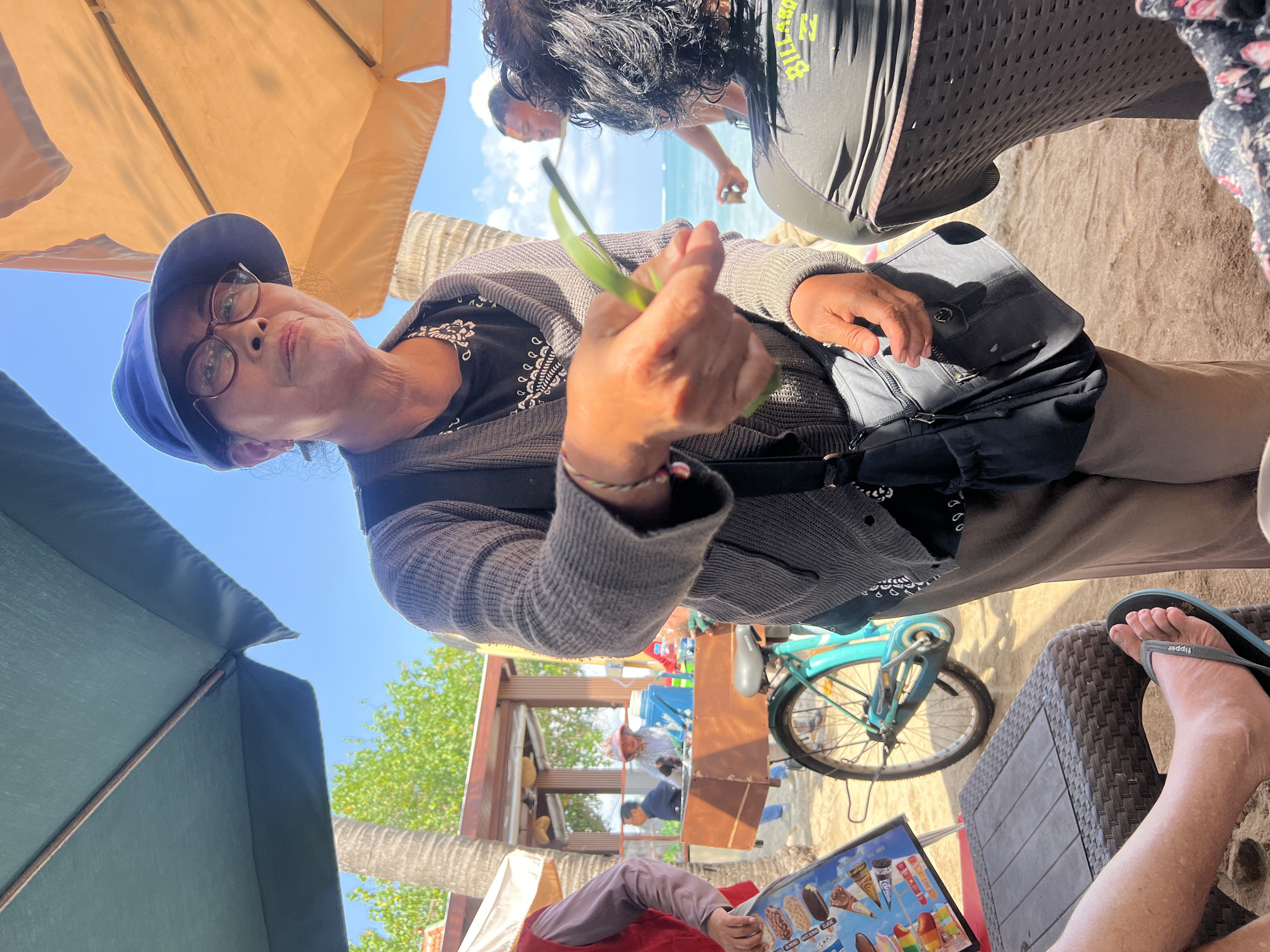 A woman wearing glasses and a blue hat is standing on a beach in Kuta, Indonesia, offering a leaf. She is wearing a gray cardigan over a black shirt and brown pants. A black bag is slung across her body. Behind her, a man is visible near the ocean, and other people are seen in the background near a food stall with a bicycle. The scene is set under beach umbrellas, with sand and water visible.