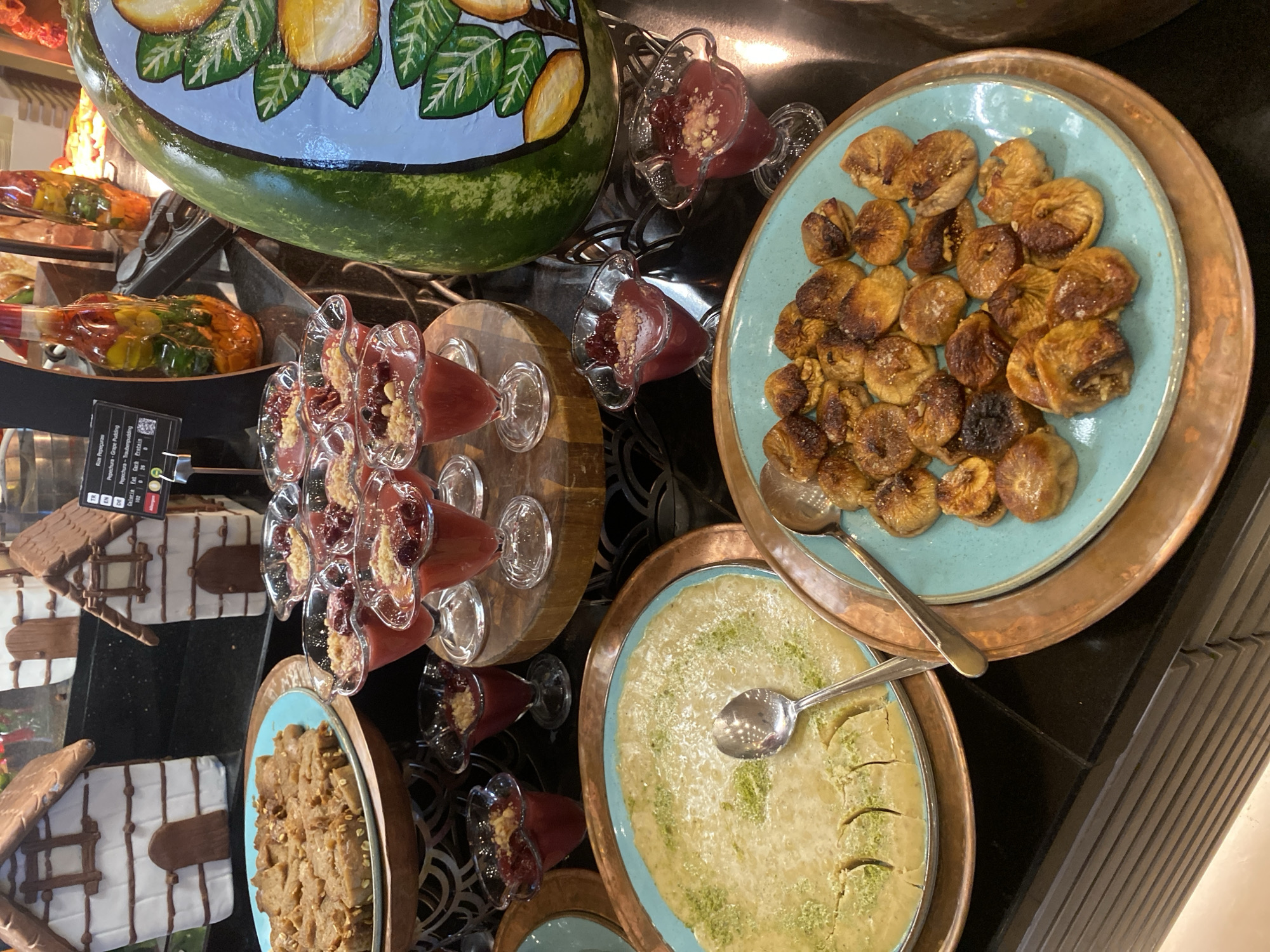 This is a close-up, eye-level shot of a dessert buffet display, likely at a hotel or restaurant in Aksu, Turkey. In the foreground, two round, copper-colored platters hold desserts on turquoise ceramic plates. The platter on the right contains a generous portion of dried figs, some of which appear to be stuffed with nuts, arranged in a somewhat haphazard but appetizing manner. A spoon rests on the figs, angled towards the right. The platter on the left holds a pale yellow, creamy dessert, possibly a pudding or custard, topped with finely chopped green nuts, likely pistachios, and cut into crescent shapes. A silver spoon is placed centrally on this dessert.
Behind these main platters, several clear glass dessert cups are arranged in rows on wooden stands. Each cup contains a reddish-pink dessert, likely a fruit compote or jelly, topped with a sprinkle of crushed nuts and possibly some small, dark fruit pieces. To the left of these cups are two miniature, gingerbread-house-like structures made of white fondant or icing with brown trim, resembling decorative elements.
Further in the background, a large, painted watermelon serves as a decorative centerpiece. It is green with painted slices of yellow fruit and green leaves. To the left of the watermelon, there are two decorative glass bottles filled with colorful liquids and what appear to be preserved fruits or vegetables. A black sign with white text is visible near the bottles, displaying information in multiple languages, including "TR," "EN," and "DE," and listing dessert names like "Rose Pépérupe" and "Pépépechura." The overall environment suggests a well-lit, indoor setting, possibly part of a larger buffet spread. No people are visible in the frame.