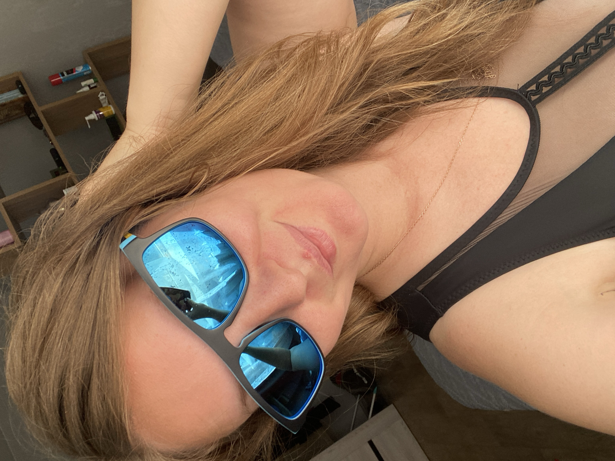 The image shows a close-up of a woman wearing sunglasses. She has long, reddish-brown hair that is partially covering her face and is resting her hand on the back of her head. She is wearing a black top with a mesh detail. The background appears to be an indoor setting with shelves holding various items, including bottles and what looks like a can of hairspray. The lighting suggests it is daytime. There are reflections in the sunglasses showing a window and possibly a person's arm. The specific location in Noviy, Russia, cannot be determined from the image.