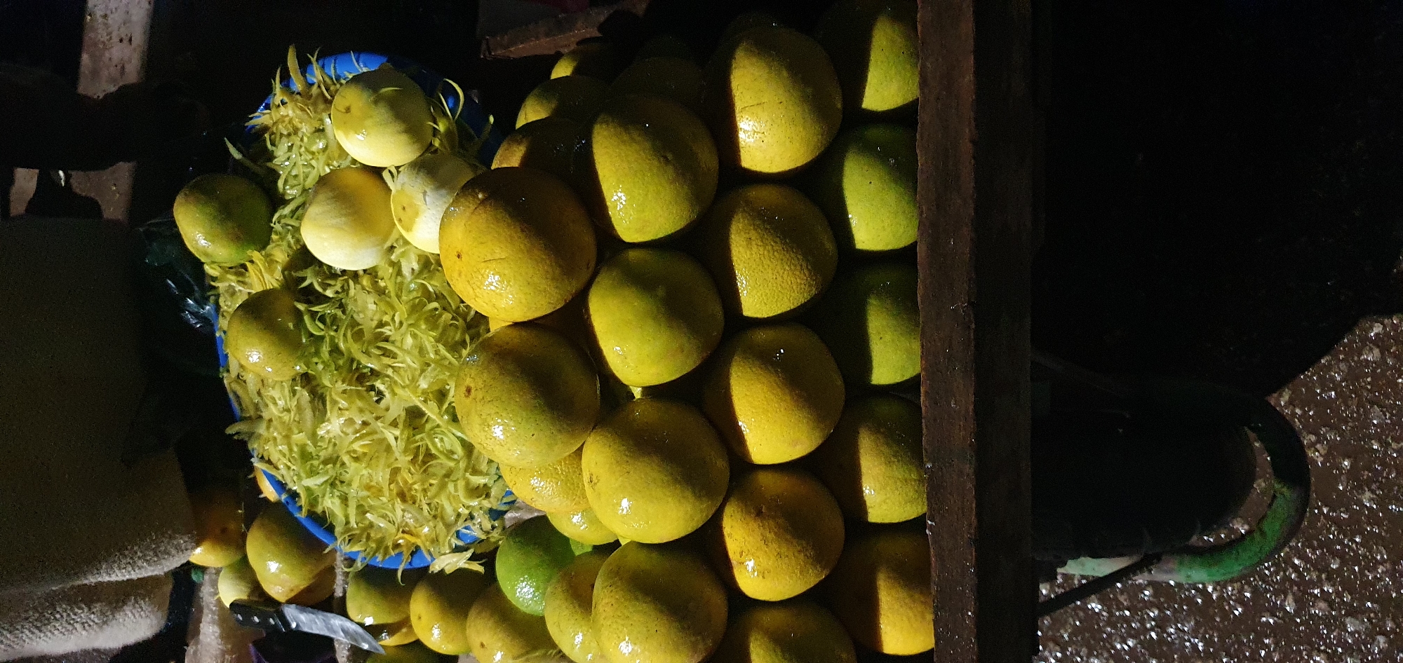 In Bukuru, Nigeria, a pyramid of wet oranges is displayed on a rustic wooden shelf. The oranges are mostly yellow-green, with some appearing more yellow. They are piled in several layers, with the top layer showing a few oranges and the layers below becoming progressively wider. Some of the oranges have water droplets on their surface, suggesting they may have been washed or sprayed. Behind the oranges, a blue plastic basin is visible, filled with shredded yellow material, possibly orange peels or zest. A knife with a dark handle and silver blade is propped against the left side of the basin. In the background, a rough, textured fabric, possibly a sack or blanket, is draped. The scene is dimly lit, creating shadows and highlighting the sheen on the oranges. The ground below the shelf appears wet and speckled with small white particles.