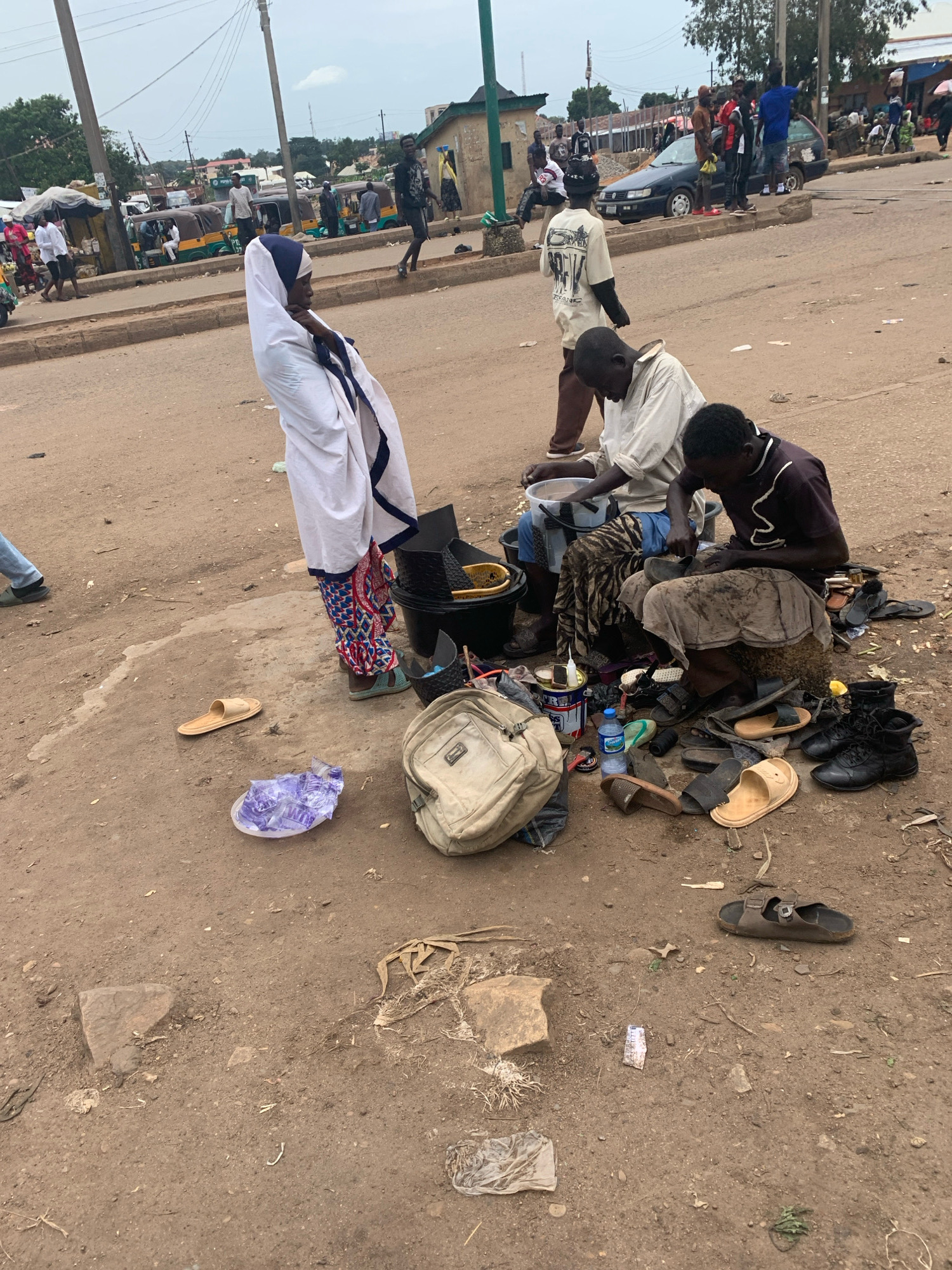 The image depicts a street scene in Bukuru, Nigeria. In the foreground, three men are engaged in shoe repair work. Two men are seated on the ground, surrounded by various footwear, tools, and containers. One man, wearing a dark t-shirt and patterned trousers, is actively working on a shoe. The other, in a light-colored shirt and brown trousers, appears to be sorting through materials. A third man stands behind them, observing.
A woman in a white and blue garment stands to the left, facing away from the camera. Scattered on the dusty ground around the shoe repair area are several pairs of sandals, a beige backpack, a plastic bag, and a water bottle. The environment is an unpaved street with buildings and a road visible in the background, suggesting a bustling commercial or residential area. Several vehicles, including what appear to be auto-rickshaws and a car, are parked or moving in the distance. Power lines are strung across the sky, typical of an urban setting. The overall impression is one of daily life and commerce taking place outdoors.