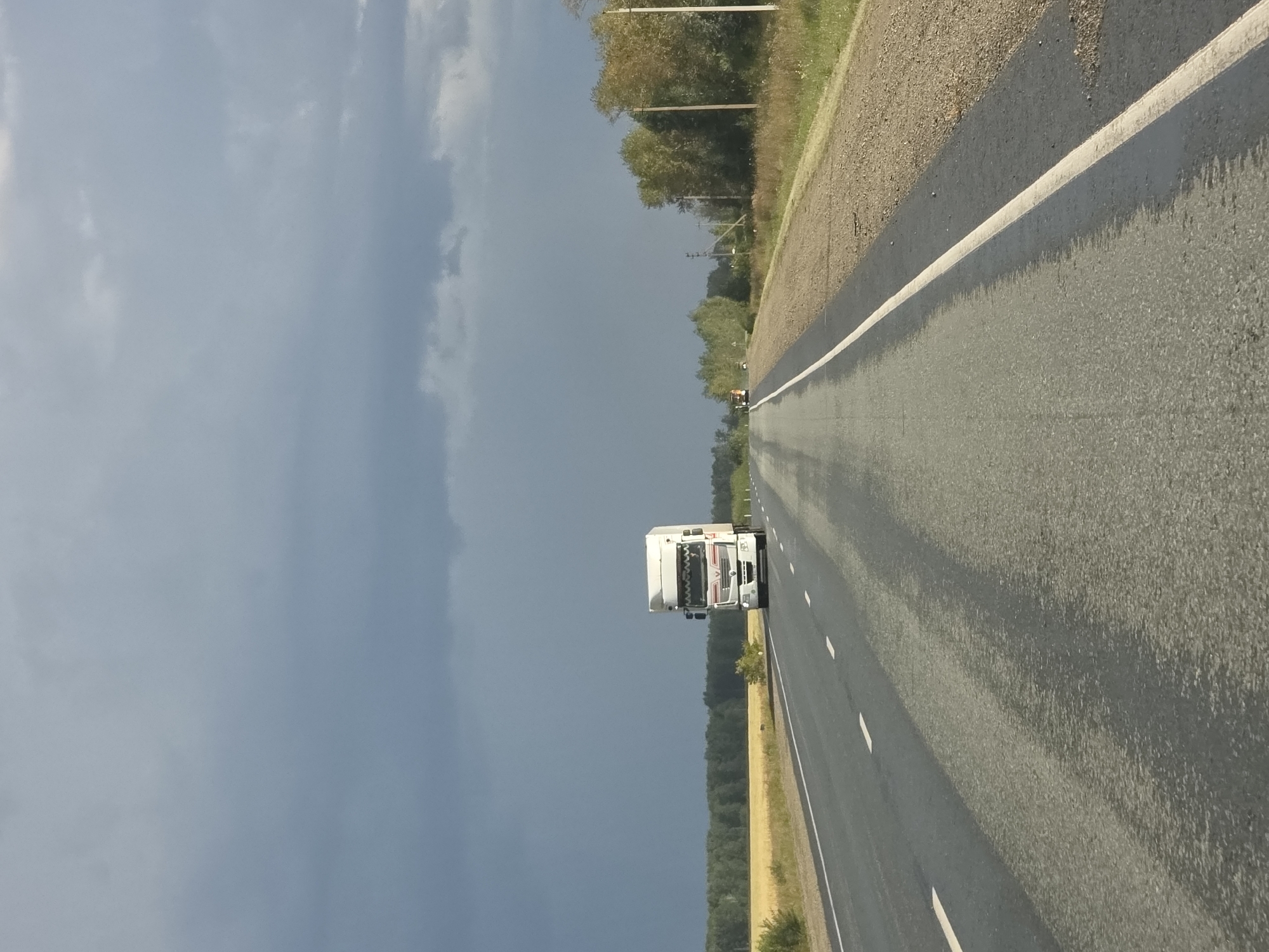 The image depicts a highway in Karsy, Russia. A white semi-truck is traveling on the asphalt road, which has white lane markings. The road is flanked by dry grass and trees on the right side and a field of yellow crops on the left. The sky above is overcast with dark, stormy clouds, suggesting impending rain. In the distance, a construction vehicle is visible on the side of the road. The scene is captured from the perspective of a moving vehicle.