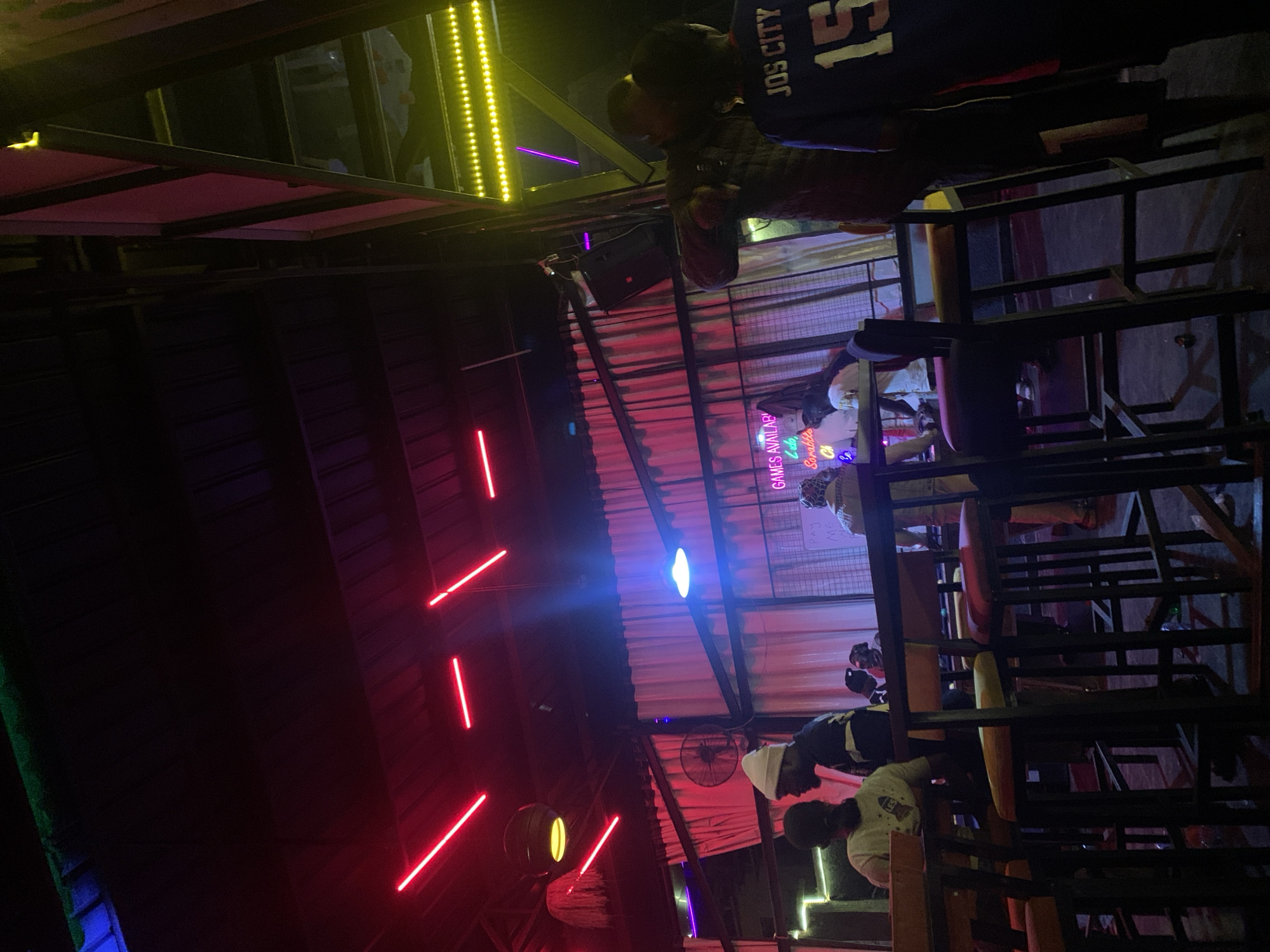 The image depicts an indoor scene with neon lighting and a dimly lit atmosphere, possibly a bar or entertainment venue. Red neon lights are prominently displayed across the ceiling. In the foreground, several bar stools and tables are visible, with a few individuals seated or standing. One person on the right wears a blue jersey with "JOS CITY 15" emblazoned on the back. In the background, a white curtain is illuminated by blue light. A neon sign in the background reads "GAMES AVAILABLE" and lists games like "Ludo, Scrabble, Chess." The setting appears to be in the city of Jos, Nigeria, specifically on or near Rukuba Road, given the "JOS CITY" text on the jersey.
