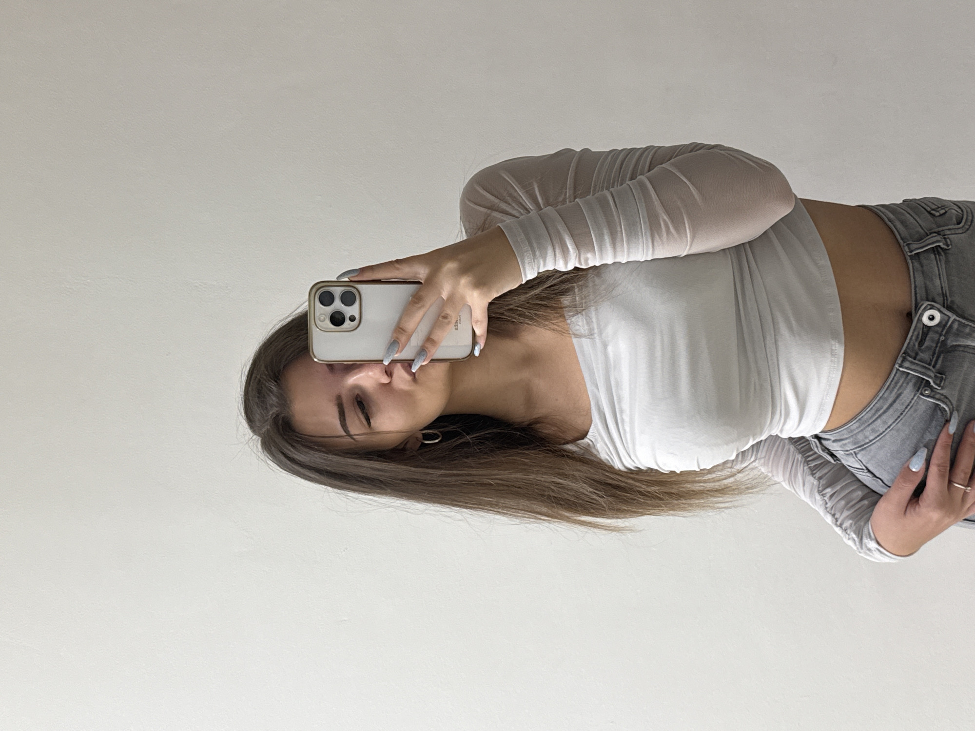 A young woman is taking a selfie in what appears to be an indoor setting. She is holding a smartphone, likely an iPhone, with her left hand, partially obscuring her face. She has long, light brown hair and is wearing a white, long-sleeved crop top and grey jeans. Her right hand is resting on her hip. The background is a plain, light-colored wall. There is nothing in the image that would indicate the location is Sofia, Bulgaria.