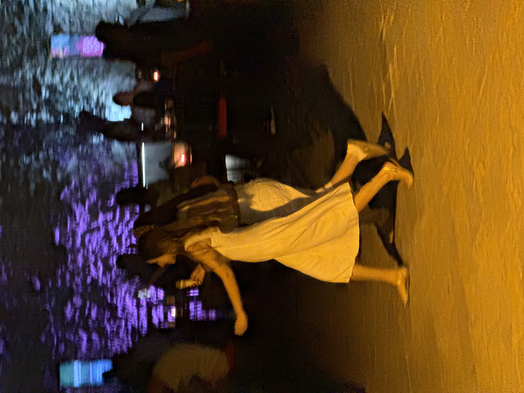 The image depicts a group of people in what appears to be an outdoor nighttime setting, possibly an event or gathering. In the foreground, two women are walking from left to right. The woman on the left is wearing a light-colored, flowing dress and holding a yellow object, possibly a drink or microphone. She has a tattoo on her arm. The woman to her right is wearing a patterned top and lighter-colored pants. Their movement suggests they are in motion, and the scene is dimly lit with prominent purple and blue lighting in the background, creating a vibrant, atmospheric effect. The environment seems to be a paved area, with indistinct figures and structures in the background, illuminated by colored lights. There are no specific identifiable landmarks that would indicate the location in Budva, Montenegro.
