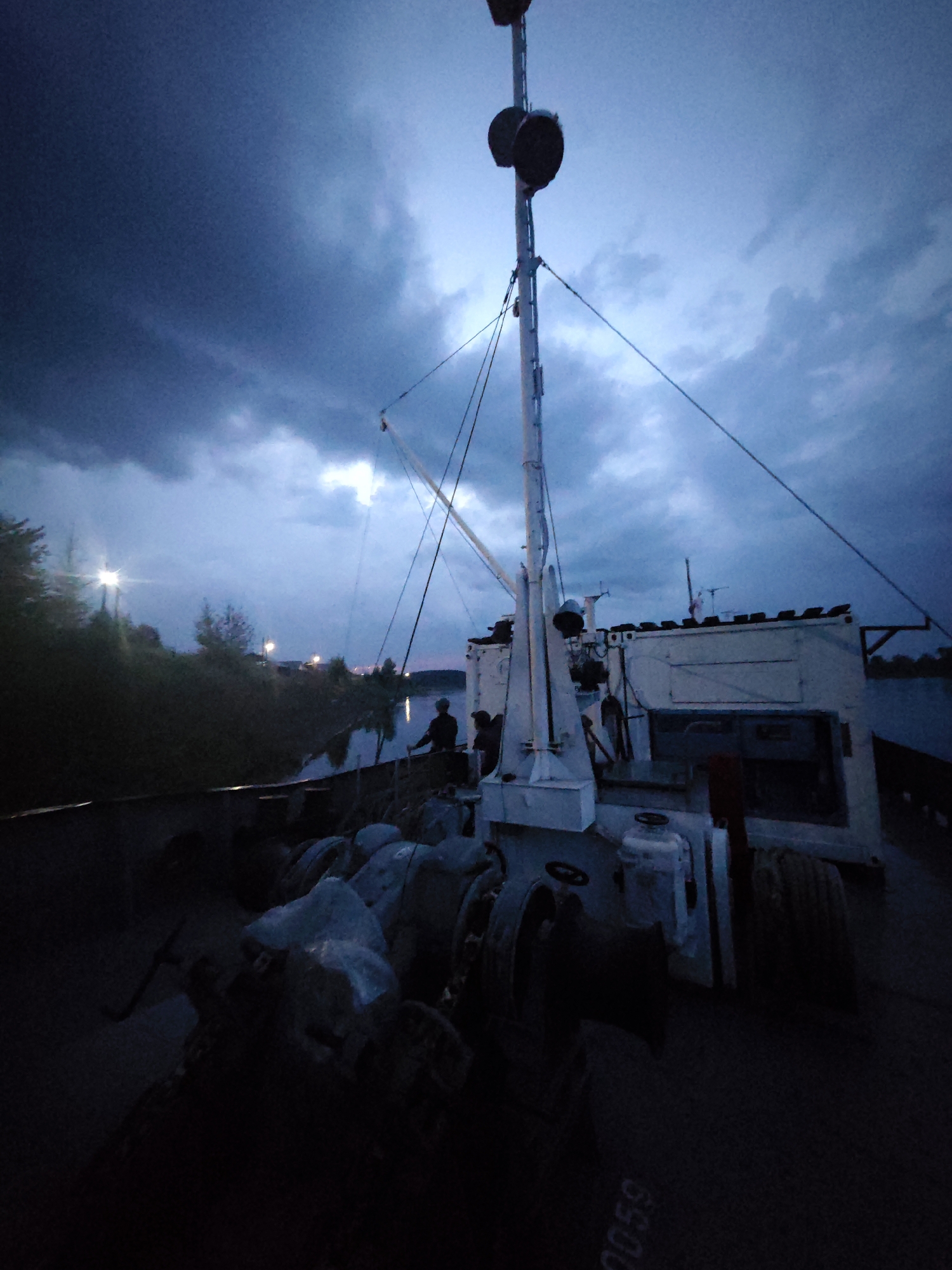 The image captures a scene on the deck of a vessel at dusk, likely near the city of Galanino, Russia. The sky is a deep blue, streaked with dark, dramatic clouds, suggesting an approaching storm or the fading light of twilight. A tall mast with two circular objects at its top dominates the upper portion of the image, supporting numerous wires and rigging.
The deck of the vessel is dimly lit, with various pieces of equipment visible. In the foreground, there are several large spools or winches, some covered with protective material. Further back on the deck, a white structure with multiple windows and a dark opening is prominent. Two individuals are silhouetted against the brighter sky in the middle ground, one appearing to be standing and looking out over the water. The dark shoreline, lined with trees and illuminated by distant lights, is visible to the left of the vessel. The water appears calm, reflecting the twilight sky. The overall atmosphere is somber and quiet, hinting at the end of a day's work or preparation for the night.