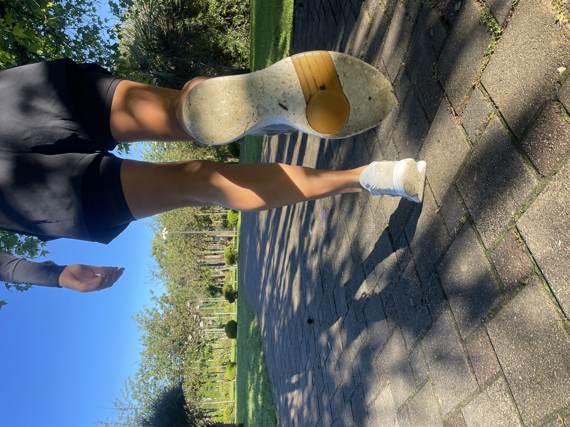 This vertically oriented image captures a low-angle view of a person's legs and feet on an outdoor paved pathway in Sochi, Russia. The person, wearing dark athletic shorts and a dark top, appears to be standing or pausing during an activity. Their right leg is slightly lifted, prominently displaying the worn and dirt-stained sole of a light-colored athletic shoe, which features the "Reebok" brand name and its delta logo, alongside a distinct yellowish-brown insert. The left foot, also clad in a matching athletic shoe, rests firmly on the patterned ground. A tanned left arm and hand are partially visible on the far left of the frame.
The setting is a well-maintained park or garden, characterized by a pathway constructed from rectangular paving stones in shades of grey and reddish-brown, with small patches of green moss or grass growing between the cracks. Beyond the path, there's a vibrant expanse of green grass bordered by diverse trees and shrubs, including leafy deciduous trees and what appear to be palm-like evergreens, consistent with Sochi's subtropical climate. The bright blue sky overhead, coupled with the long, distinct shadows cast by the trees across the pathway, indicates a sunny morning or late afternoon. The image conveys a sense of outdoor activity on a clear, pleasant day.