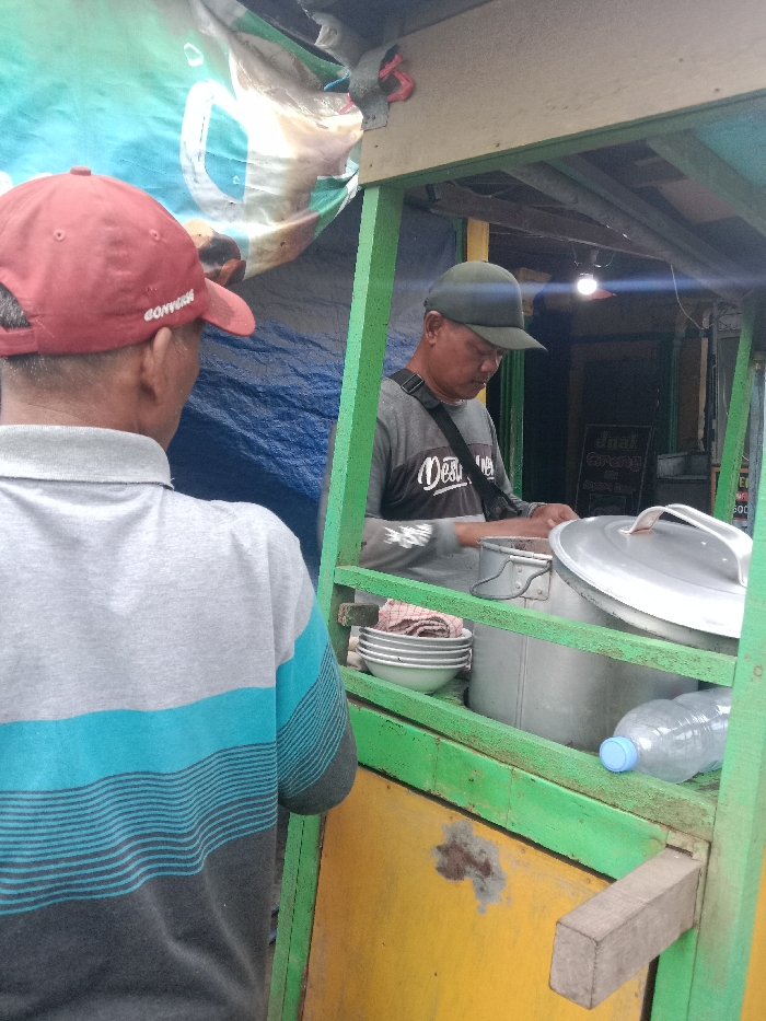Two men stand at a food cart. One wears a red baseball cap and a gray and blue striped shirt, facing the cart. The vendor inside the green-framed cart wears a dark baseball cap and a gray shirt with black lettering. He is preparing food with metal pots and bowls arranged on a shelf. A plastic water bottle sits on the bottom shelf. A sign is visible in the background.