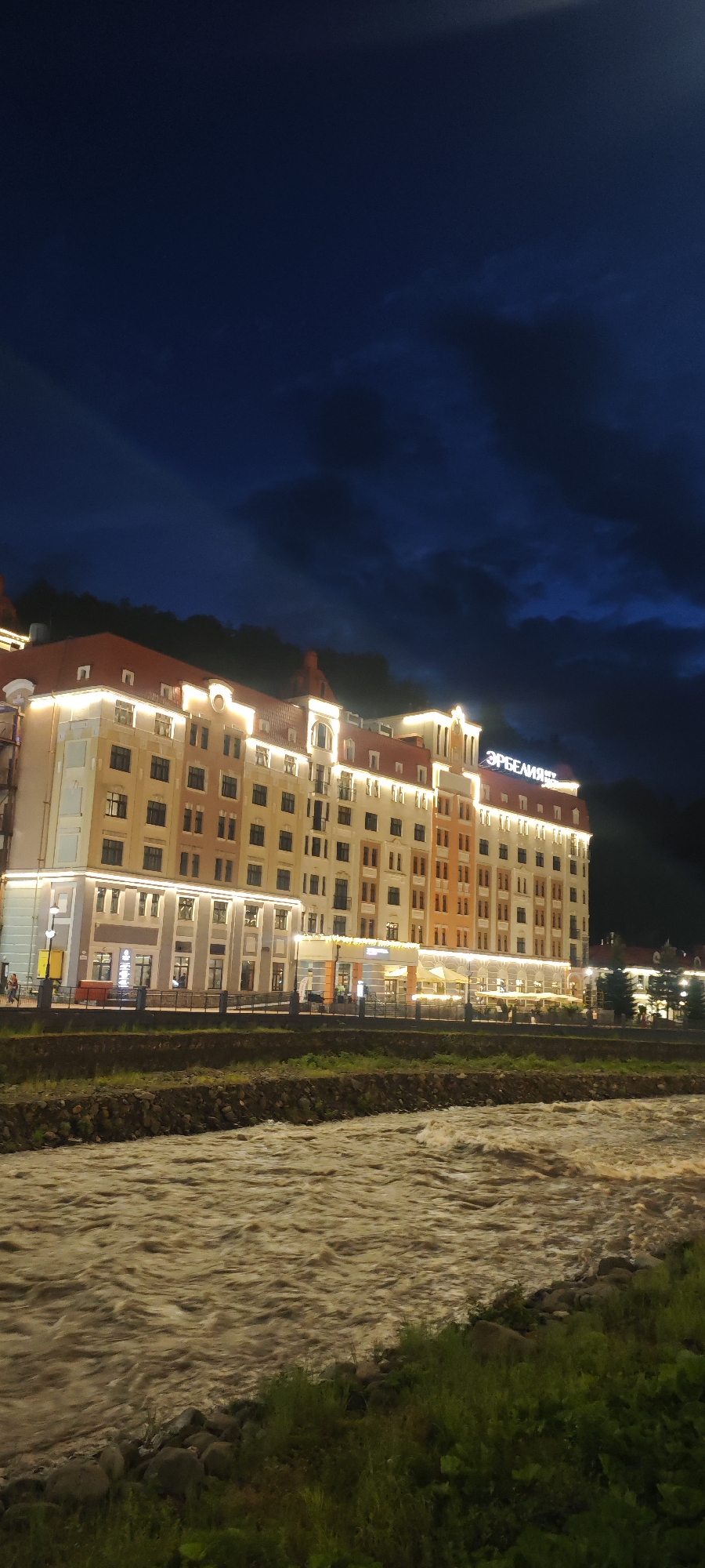 The image presents a nighttime view of a building, identified by its sign as "Erbelia," situated in the city of Estosadok, Russia. The structure is a multi-story building with a red roof and illuminated windows. The building's façade is a mix of beige and orange. The name "Erbelia" is displayed in illuminated Cyrillic characters at the top of the building.
In the foreground, a river flows rapidly with muddy-brown water. The riverbank is covered with grass and rocks.
The sky is dark blue with some cloud cover. Mountain silhouettes are visible in the background. The lighting suggests that the image was taken at night. There are people walking near the building along the sidewalk.