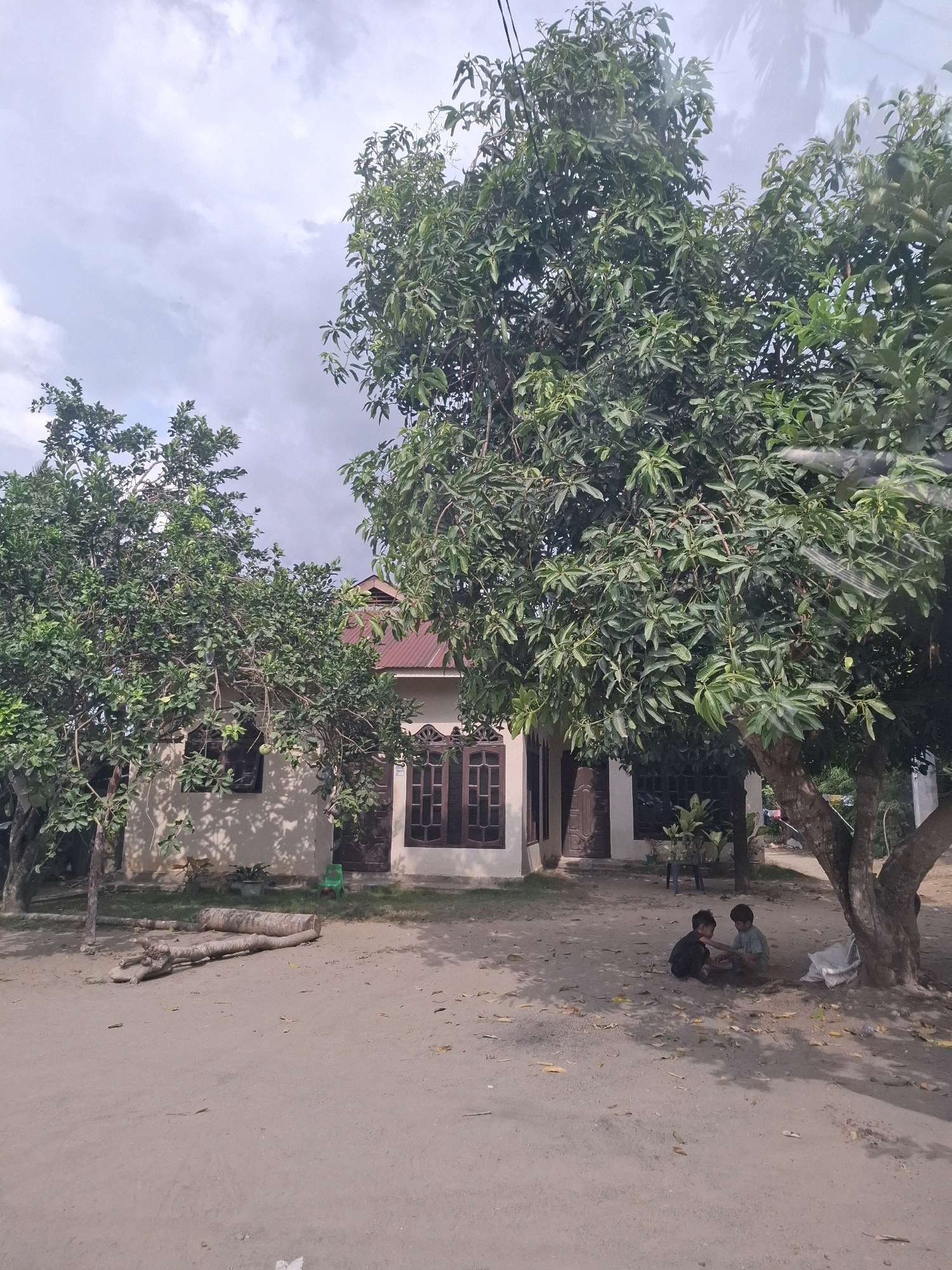 In the city of Awegeutahpaya, Indonesia, two young children are seated on the dirt ground in front of a single-story house with a reddish-brown metal roof and cream-colored walls. The house has dark-paned windows and doors. A large tree with dense green foliage stands to the right of the house, casting shadows on the ground. Two fallen logs are positioned in the dirt near the left side of the house. The sky above is overcast with scattered clouds.