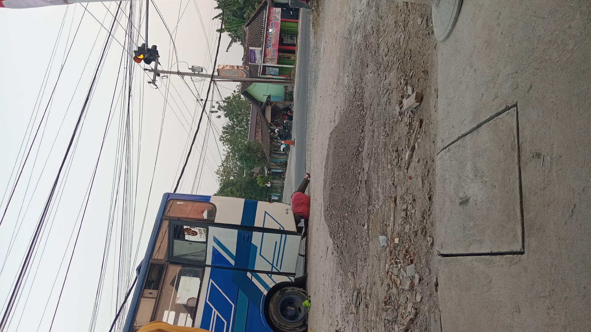 The image depicts a roadside scene in Trembulrejo, Indonesia, where a person in a red shirt and dark pants is lying on their back underneath a blue and white bus. The bus is parked on the side of a paved road, and the person appears to be working on the underside of the vehicle. The environment is dusty, with piles of gravel and construction debris scattered around. In the background, there are several buildings, including shops and houses, as well as utility poles with numerous overhead wires. A traffic light with a yellow signal is visible in the distance. The sky is overcast, suggesting it might be early morning or late afternoon.