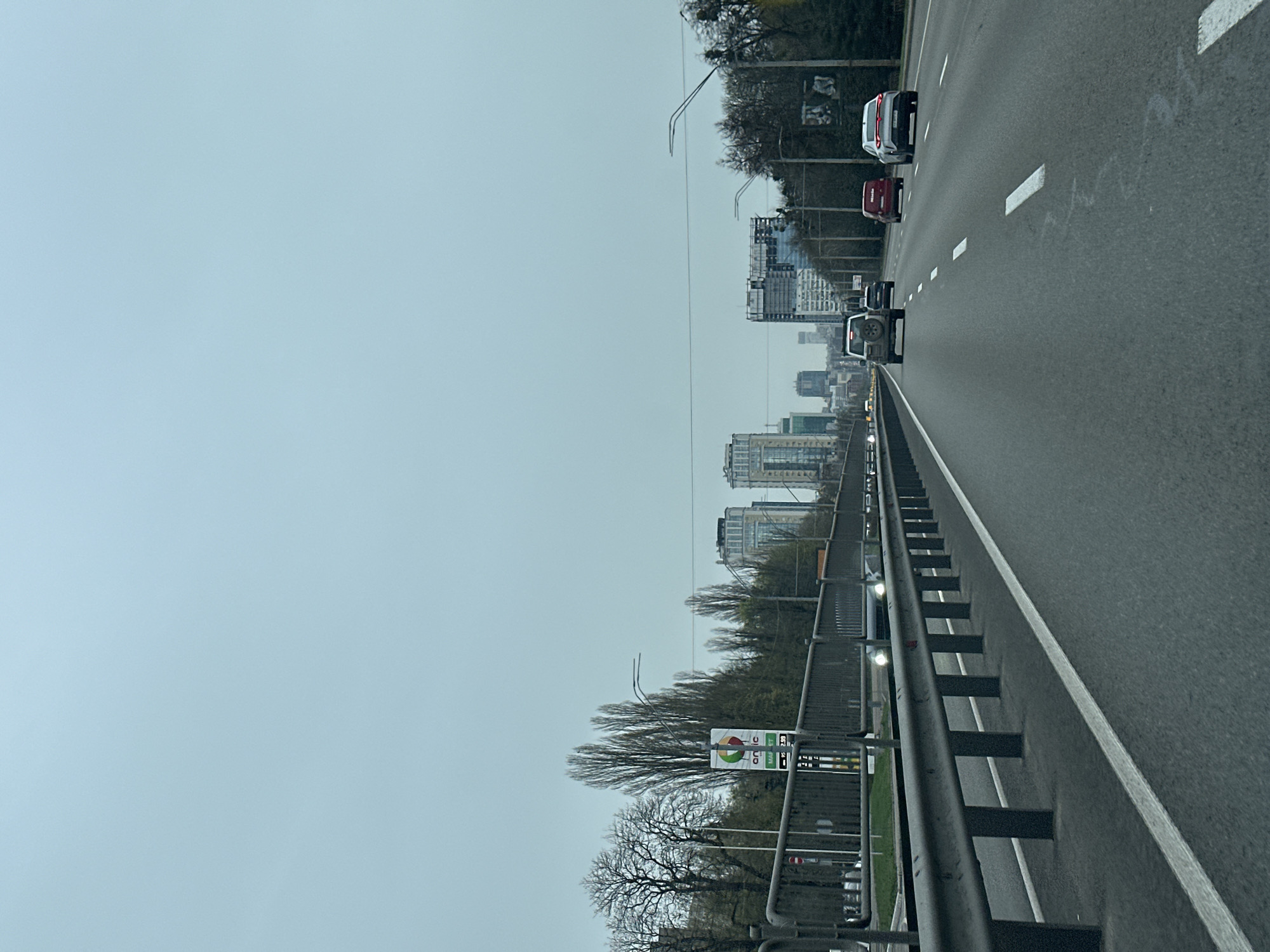 The image depicts a multi-lane highway in Kyiv, Ukraine. The view is from a moving vehicle, showing traffic flowing in the same direction. Several cars are visible, including a white SUV and a red car. On the side of the highway, a metal guardrail separates the road from trees and buildings. Distant high-rise buildings and infrastructure are visible in the background, obscured by a hazy or overcast sky. A roadside sign is also visible.