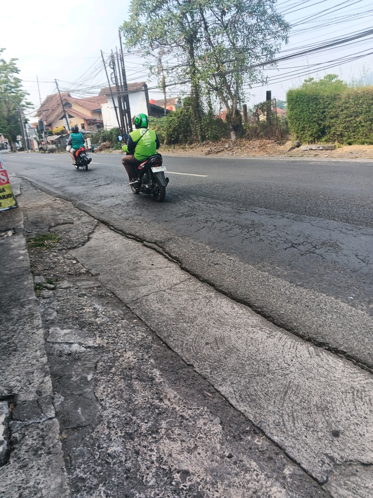 A man wearing a green helmet and a bright green vest with the Gojek logo is riding a motorcycle on a paved road in Ungaran, Indonesia. He is in the right lane, and another person on a motorcycle is in the left lane ahead of him. The road appears to be in moderate condition with some cracks. On the left side of the road, there is a concrete curb and sidewalk. Buildings and trees are visible in the background, along with utility poles and wires. The sky is overcast. The scene depicts a common mode of transportation and delivery service in the area.