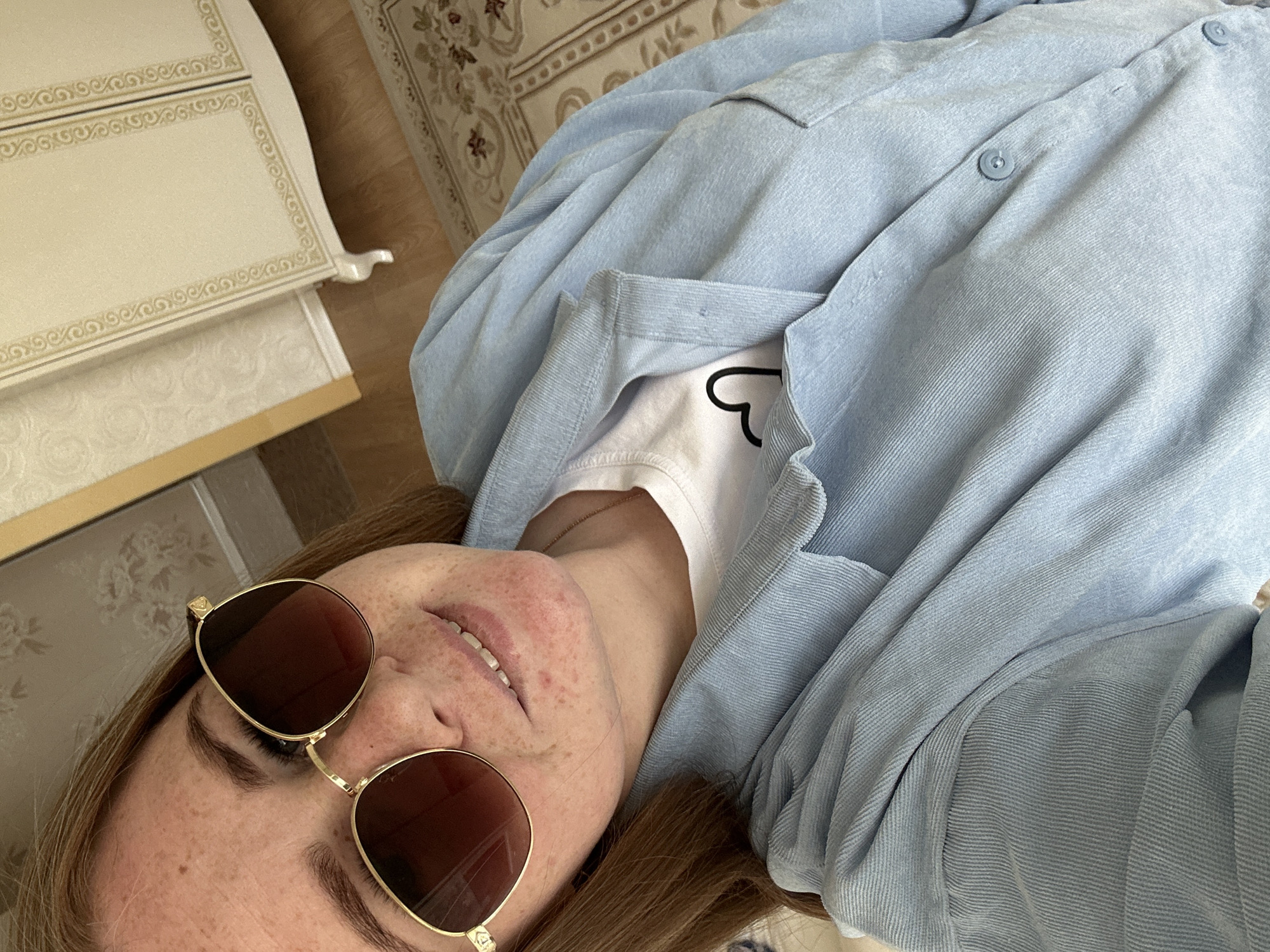 A woman with freckles is lying down, wearing gold-rimmed sunglasses with brown lenses, a white t-shirt with a partial black heart graphic, and a light blue button-down shirt. She is positioned diagonally in the frame. To the left is a white and gold patterned dresser. A rug with a floral pattern is partially visible in the upper-center background. The location is Chaikovsky, Russia.