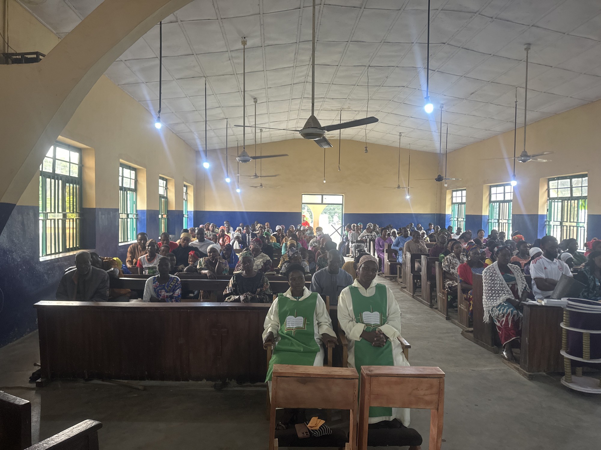 The image shows the interior of a church in Akwanga, Nigeria. The church has a high, vaulted ceiling with several ceiling fans. The walls are painted yellow and blue, and the windows are covered with green bars. Rows of wooden pews fill the church, and the floor is made of concrete.
In the foreground, two individuals, likely clergy members, are seated in chairs. They are wearing green and white vestments with a book symbol on them. In the pews behind them, a congregation of men, women, and children are seated. Many of the women are wearing headscarves. The people appear to be attentive, facing towards the front of the church.
In the background, more of the congregation can be seen, and through a doorway, glimpses of greenery are visible, suggesting an outdoor area beyond the church. The overall atmosphere appears to be one of a religious service in progress.