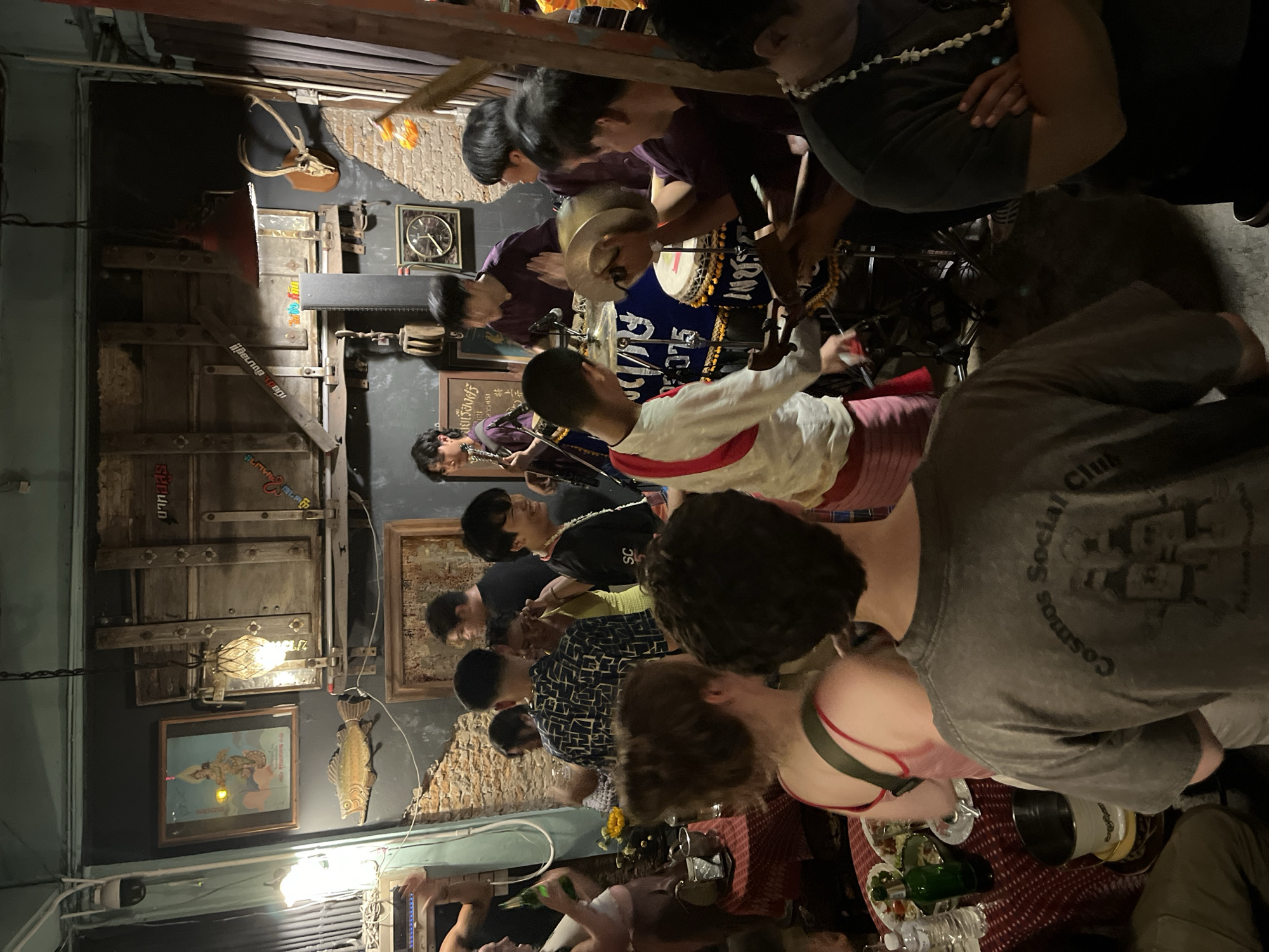 The image shows a group of people gathered in a dimly lit indoor space, likely a bar or restaurant in Chiang Mai City Municipality, Thailand. A band performs, with members playing instruments like drums and stringed instruments. Patrons are seated at tables, some with food and drinks. The decor includes vintage items such as clocks, pictures, and a decorative fish on the walls. The environment appears to be a casual and lively social setting.