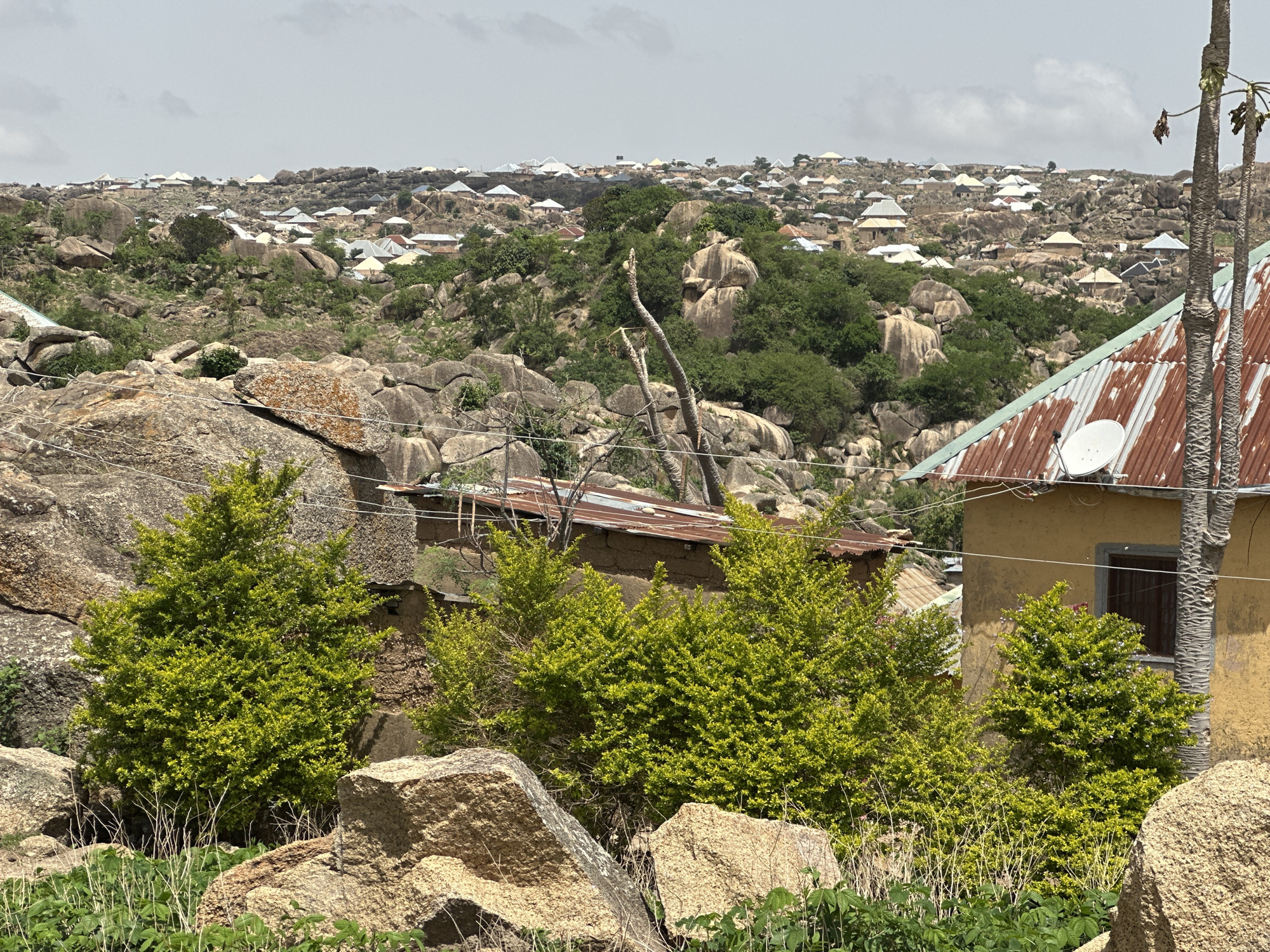 The image shows a view of a rocky, hilly landscape in the city of Jos, Nigeria, dotted with houses and trees. In the foreground are large rocks and bushes. A yellow building with a corrugated metal roof and a satellite dish is visible on the right. A smaller structure with a similar roof is partially obscured by the bushes. A line of utility wires spans the scene. In the background, numerous houses with light-colored roofs are spread across the terrain. The sky is overcast with light clouds.