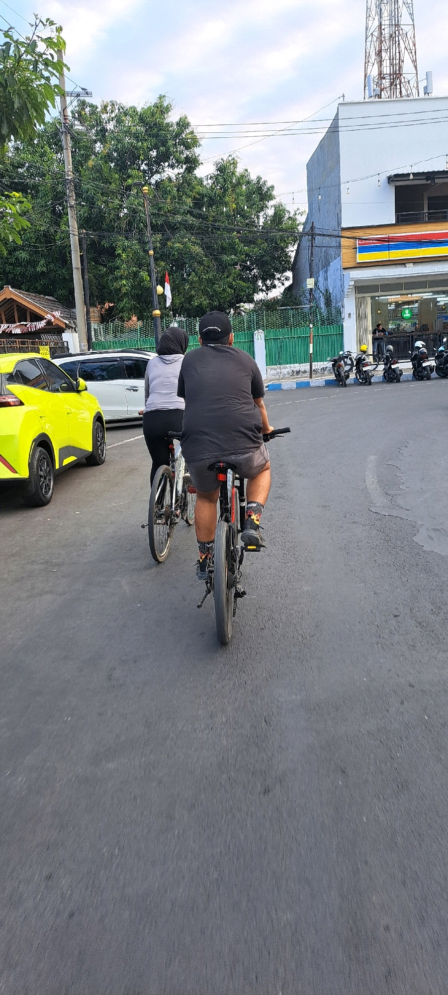 Two people are riding bicycles on a paved road in Pasuruan, Indonesia. The person in front is wearing a grey hoodie and a black hijab. The person behind them is wearing a dark grey t-shirt and grey shorts. Both individuals are facing away from the camera. To the left of the cyclists is a bright yellow car. In the background, there is a building with a white facade and a blue stripe, appearing to be a commercial establishment, possibly a convenience store, with motorcycles parked in front. Overhead, there are power lines and a telecommunications tower. Lush green trees and foliage are visible behind the cyclists and to the left. The sky is overcast with some clouds. The overall setting suggests an urban or suburban environment.