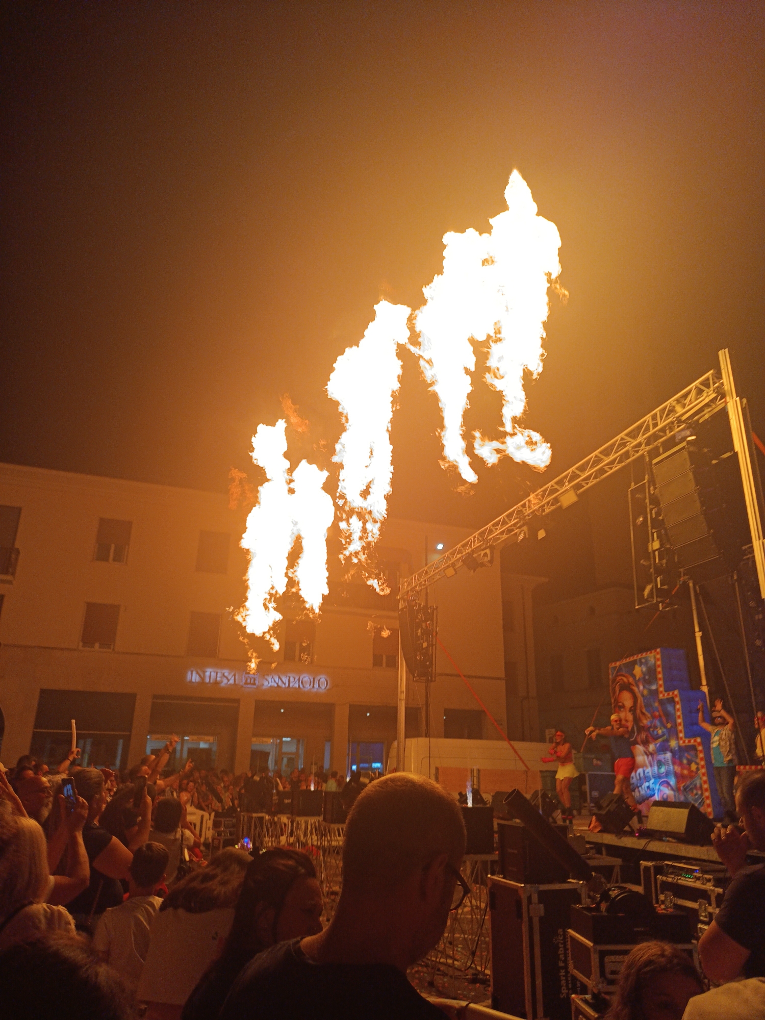 This is a photograph taken at night during an outdoor event in Viadana, Italy. The sky is dark and illuminated by bright orange and white flames erupting from a stage or structure on the right side of the image. These flames are the primary light source, casting a warm glow on the surrounding buildings and the crowd of people gathered below.
In the background, a light-colored building with windows and balconies is visible. A prominent sign on this building reads "INTESA SANPAOLO". To the right of the building, a stage setup with scaffolding, large speakers, and lighting equipment is apparent. A colorful backdrop featuring a portrait of a woman is part of the stage design.
The foreground and midground are filled with a crowd of people, many of whom are looking towards the stage and the flames. Some individuals are holding up their phones, presumably to take pictures or videos. The atmosphere appears to be festive and energetic, with the spectacle of the flames likely being a focal point of the event. The overall scene suggests a live performance or a public celebration.