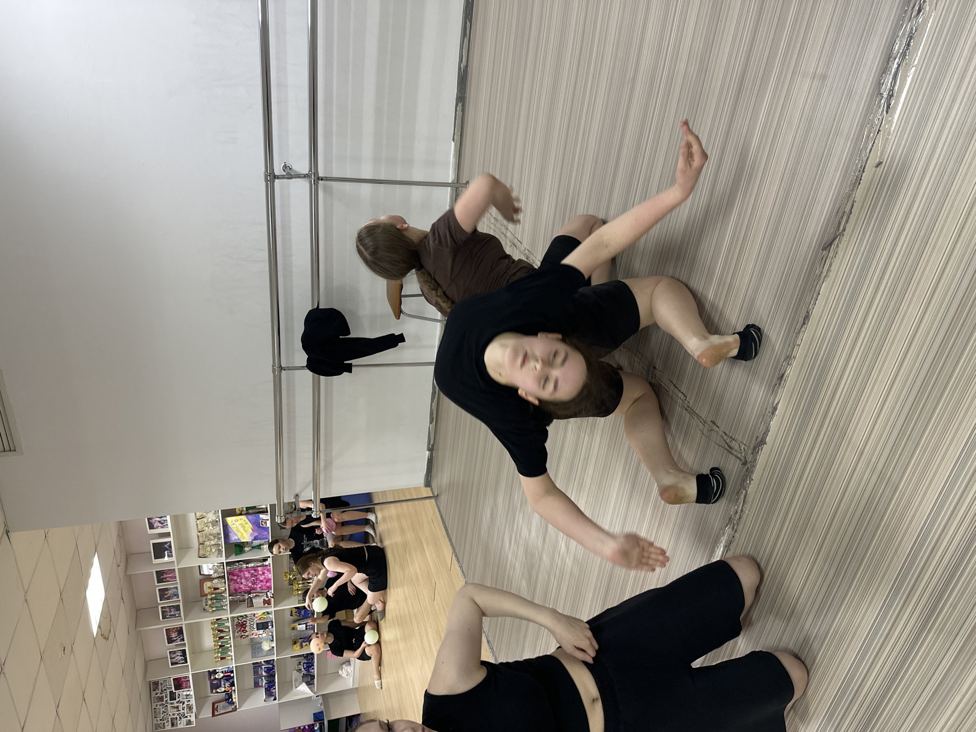 The image captures an indoor scene in what appears to be a dance or fitness studio in Votkinsk, Russia. Several young people, likely teenagers or young adults, are engaged in a movement activity, possibly contemporary dance, stretching, or floor work.
In the foreground, a person wearing a black t-shirt and black shorts lies on their back/side on a light-colored, striped floor, looking towards the right. Another individual, also in black activewear, is positioned further back, seemingly in a dynamic pose on the floor with arms extended. Behind them, a third person, dressed in a brown t-shirt and black shorts, sits or kneels, looking towards the plain white wall. This wall features a metallic ballet barre.
A large mirror covers a significant portion of the right side of the room, reflecting additional participants. These reflected individuals are also in activewear, and some appear to be holding small, light-colored balls, suggesting activities like Pilates or specific stretching exercises.
On the far left, a multi-tiered shelving unit is visible, densely packed with numerous trophies, framed certificates, and photographs, indicating a history of awards or achievements for the studio or its members. A black hoodie or jacket hangs on the ballet barre.
The setting is well-lit, suggesting an ongoing class or practice session. The participants appear focused on their movements. No specific text is clearly legible on any of the awards or decorations.
