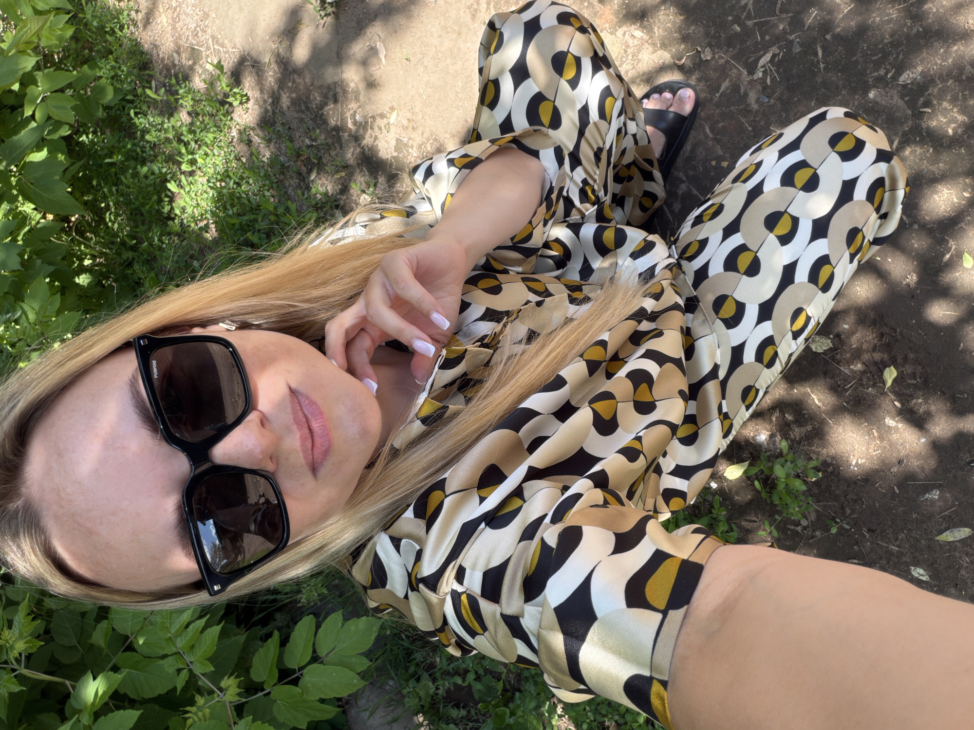 The image depicts a fair-skinned female individual with long, straight blonde hair, wearing large black square-framed sunglasses branded "Polaroid." Her left hand, featuring a white French manicure, is raised with fingers lightly touching her jawline. She is dressed in a two-piece outfit, likely a shirt and pants, made of a silky material with a repetitive geometric pattern of concentric circles in white, beige, black, and mustard yellow. Black open-toed sandals are visible on her feet. The individual is positioned outdoors, appearing to take a selfie. The background features dense green foliage on the left and upper left, and a dirt ground with sparse green weeds and scattered dry leaves on the right and foreground. Dappled sunlight creates distinct shadows across the scene. The precise location in the city of Votkinsk, Russia, cannot be forensically determined from the visual content of the image.