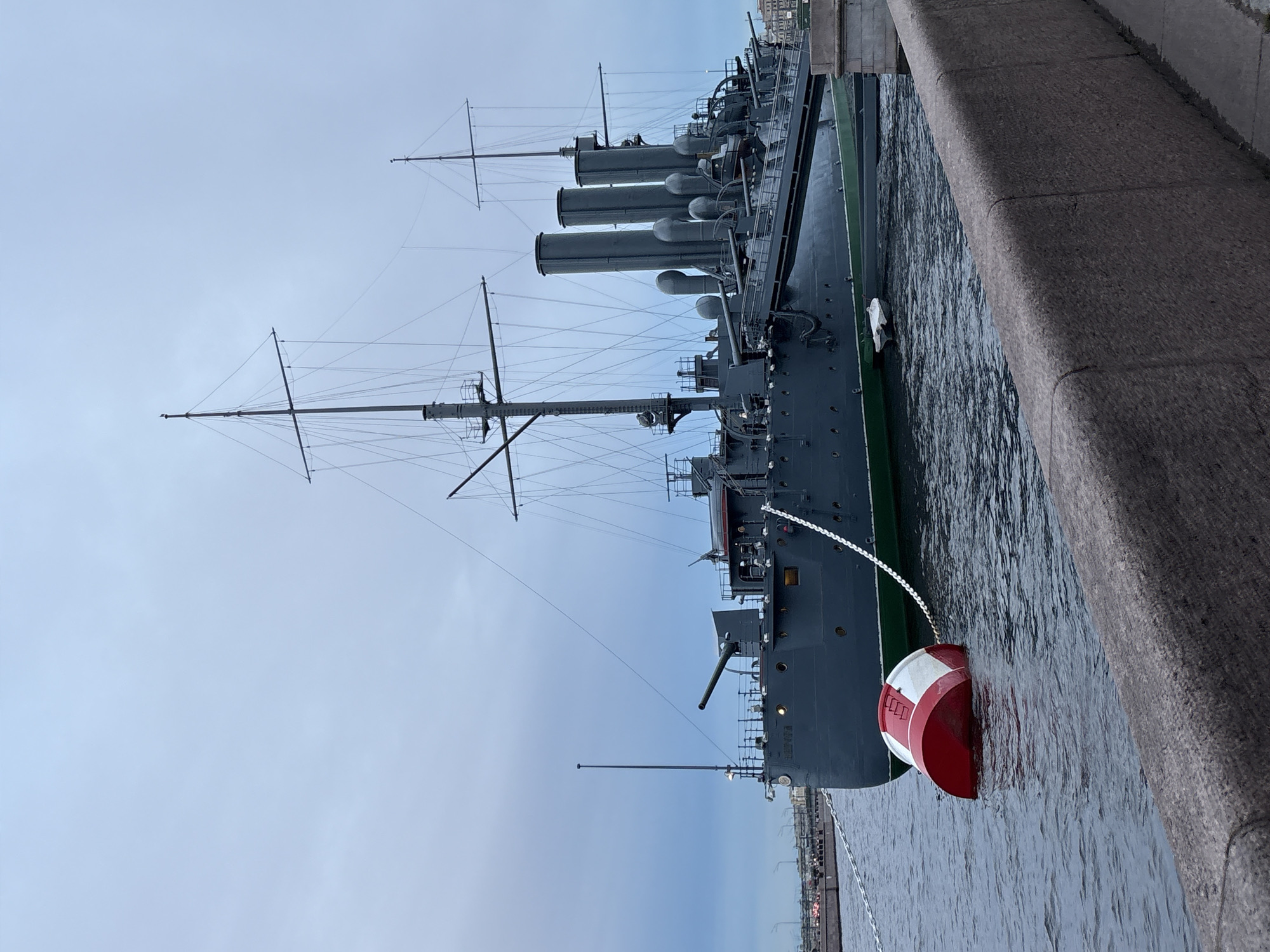 The image shows a side view of a large, dark gray warship docked next to a stone embankment. The ship has multiple smokestacks and masts, suggesting it is an older vessel. A red and white buoy is tethered to the ship with a chain, floating in the water. The water appears choppy, and the sky is overcast and gray, indicating a cloudy day. The scene is likely a port or harbor in Saint Petersburg, Russia, given the architectural style of the embankment and the type of ship. There are no people visible in the image, and the overall mood is somber and still, reflecting the overcast weather and the historical nature of the warship. The prominent text on the buoy is illegible due to the image resolution.