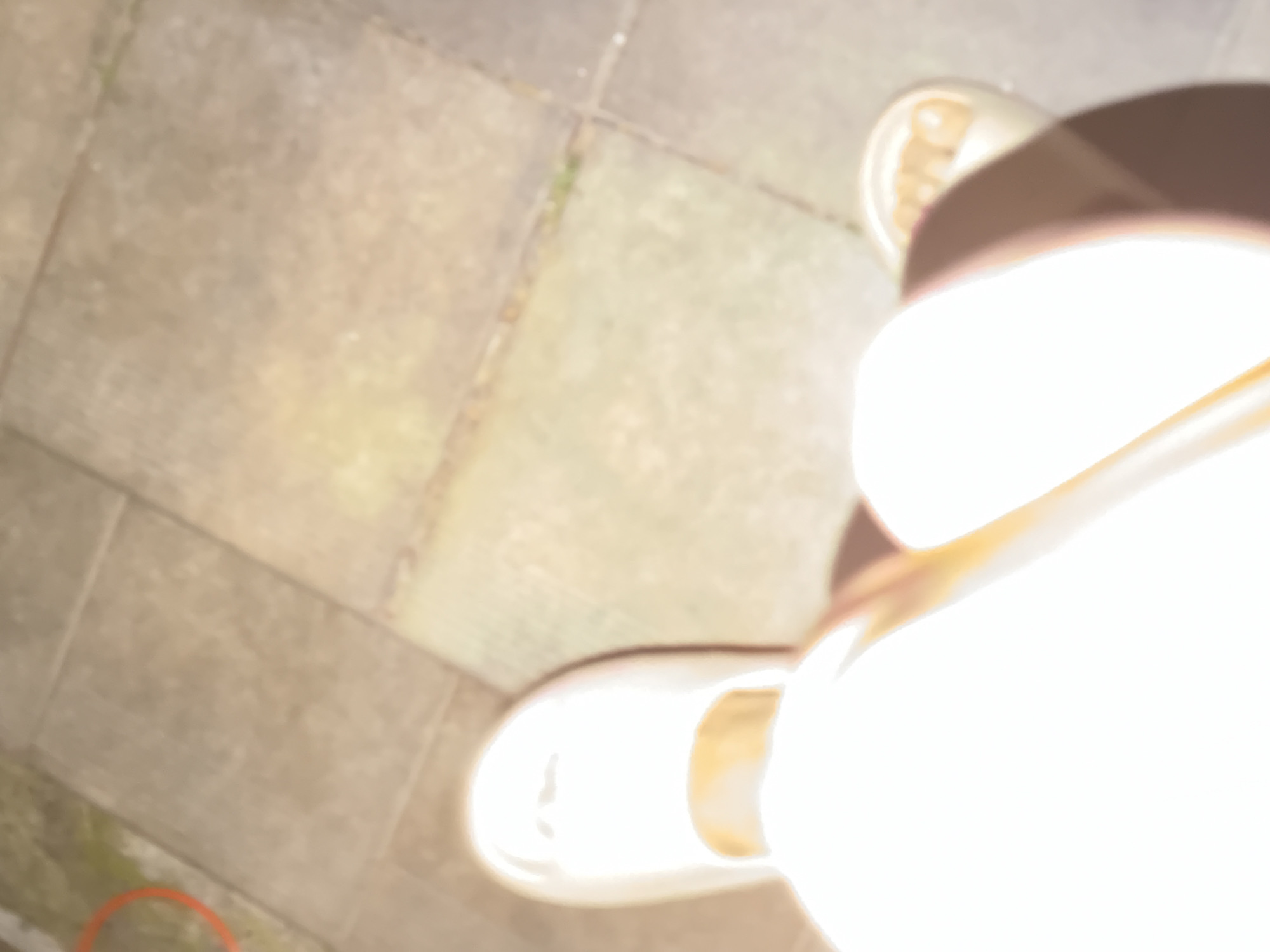 The image shows a pair of feet wearing light-colored slip-on shoes. The feet are positioned on a tiled outdoor surface, possibly a patio or walkway. The lighting suggests it is daytime, with a bright glare casting shadows. The scene is blurred, making specific details difficult to discern. There is no indication of the city of Bila Tserkva, Ukraine, nor any other contextual information that could place the image in a specific location.