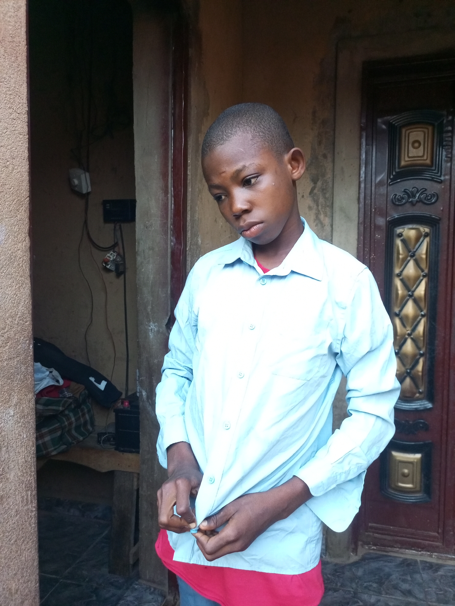 A young boy, with closely cropped hair and a serious expression, stands in a doorway. He is wearing a light blue button-down shirt over a red garment. His hands are in the process of buttoning his shirt. The setting appears to be outdoors, in front of a building with a textured, light brown wall on the left and a dark brown, ornate door on the right. Behind the boy, an open doorway reveals a glimpse of an interior space with electrical wiring and some stacked items, including fabric with a plaid pattern and dark clothing. The lighting suggests it could be daytime, with some shadows indicating it's not direct overhead sun. The overall mood is calm, with the boy focused on his task. The image was taken in Tempure, Nigeria. There is no visible text in the image.
