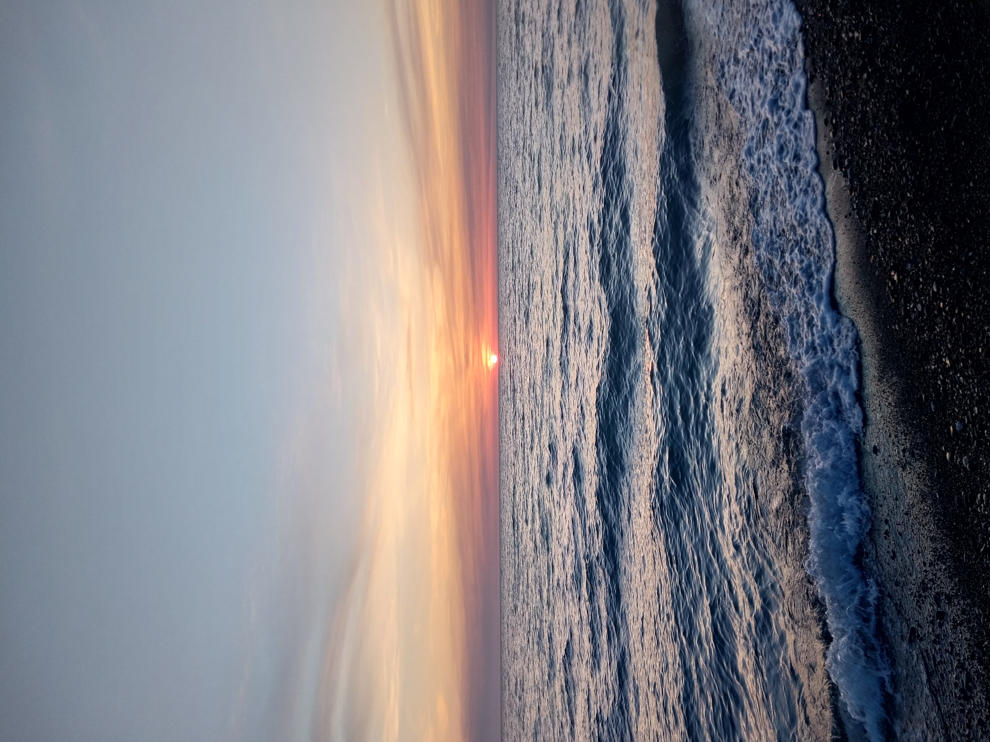 The image depicts a sunset over the Black Sea, near Sochi, Russia. The left side of the image shows the sky transitioning from light blue to orange and pink hues, indicating the setting sun. The sun is visible on the horizon. The right side shows the sea with visible waves and surf. The foreground shows a dark rocky beach. There are no people visible in the frame.