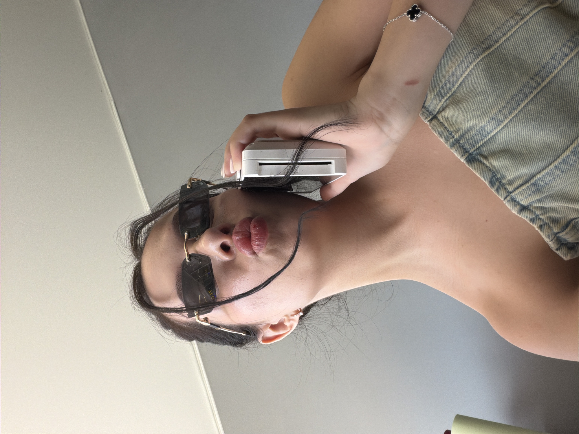 The media file is a close-up, rotated photograph featuring a young person's face. The person is wearing dark-rimmed sunglasses and has their lips pursed as if to kiss. Their dark hair falls around their face, with some strands obscuring the view of their sunglasses. They are holding a small, white electronic device up to their face. The background is a neutral gray wall, with a lighter, possibly white, surface in the upper left corner, suggesting an indoor setting. The lighting appears to be soft and diffused, indicating it's likely daytime. The person is wearing a light-colored denim garment, visible on their right shoulder and arm, which has visible stitching. A thin silver bracelet with a four-leaf clover charm is visible on their left wrist. There is a small reddish mark on the skin of their left forearm. No visible text is present in the image. The overall impression is of a selfie taken in a casual, personal setting. The location context of Kamianske, Ukraine, is noted but not visually discernible from the image itself.