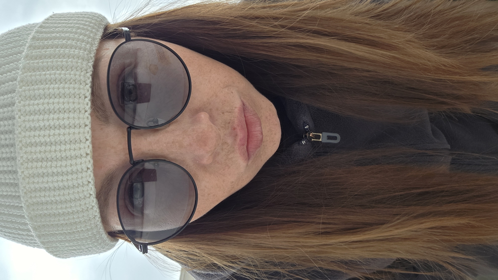 A person with fair skin and freckles is wearing a white knitted beanie and round, dark sunglasses. The person has reddish-brown hair, and the camera angle is a close-up portrait. The person is wearing a dark-colored jacket, with a zipper visible at the collar. The sky is visible in the background, suggesting an outdoor setting. The city location is Unknown, Unknown.
