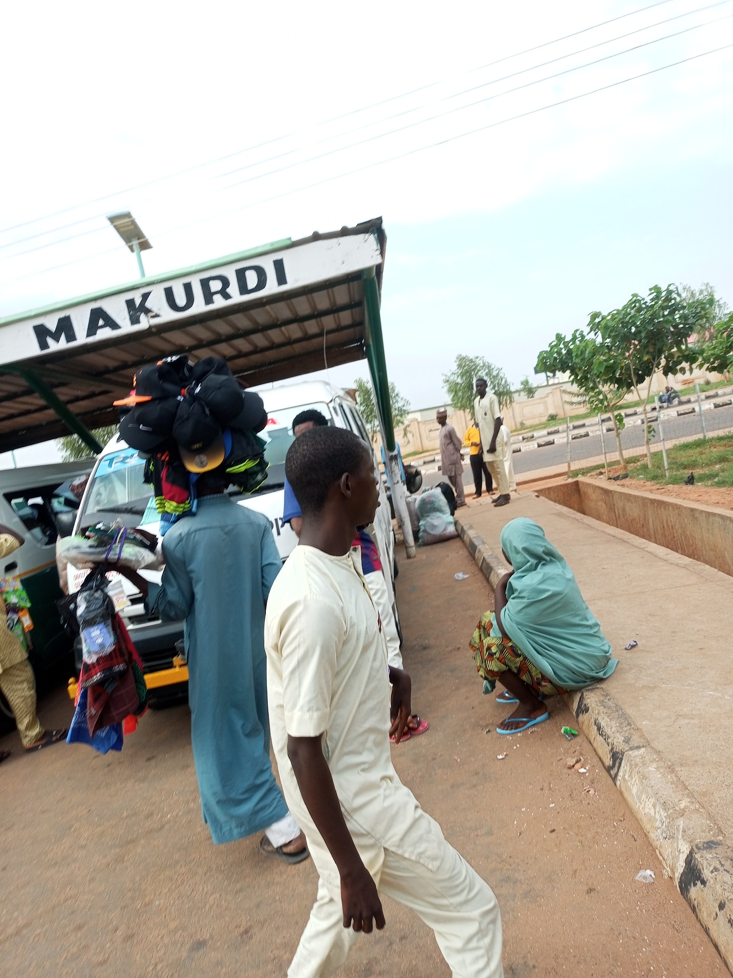 The image depicts a street scene in Gombe, Nigeria, likely a bus park or a busy roadside market. The sign "MAKURDI" is visible, suggesting a connection to the Makurdi transport route or destination.
In the foreground, a man dressed in a light-colored kaftan is walking away from the camera, his back partially visible. To his left, another man in a teal robe is carrying a large stack of black caps and some fabric items on his head, with a basket of goods in his hand. He is standing near a white van, which appears to be a commercial vehicle. Further back, several other individuals are present, including a man in a yellow shirt and a man in a brown kaftan.
To the right of the scene, a woman is seated on the curb, wearing a teal headscarf and a patterned skirt. She is looking towards the road. The environment includes a paved walkway, a road with vehicles in the background, and some trees and vegetation. The ground is a mixture of dirt and concrete. The overall impression is one of a lively, bustling urban environment.