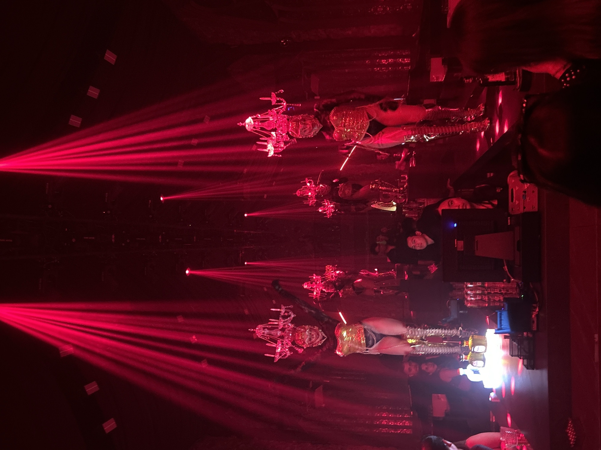 The image depicts a performance taking place in a dimly lit indoor venue, possibly a nightclub or stage show, in North Kuta, Indonesia. The dominant color is a deep red, emanating from dramatic stage lighting that casts strong beams across the scene. In the foreground, several individuals are partially visible, some seated and others standing. The main focus of the image is on two performers on stage, positioned towards the center. These performers are elaborately costumed, wearing outfits that appear to be metallic or reflective gold, featuring high boots and decorative headdresses resembling chandeliers. Their poses suggest they are actively engaged in a performance. The background shows more stage elements, including lighting fixtures and possibly curtains or textured walls. The overall atmosphere is one of entertainment and possibly theatricality, amplified by the intense red lighting and the striking costumes of the performers.