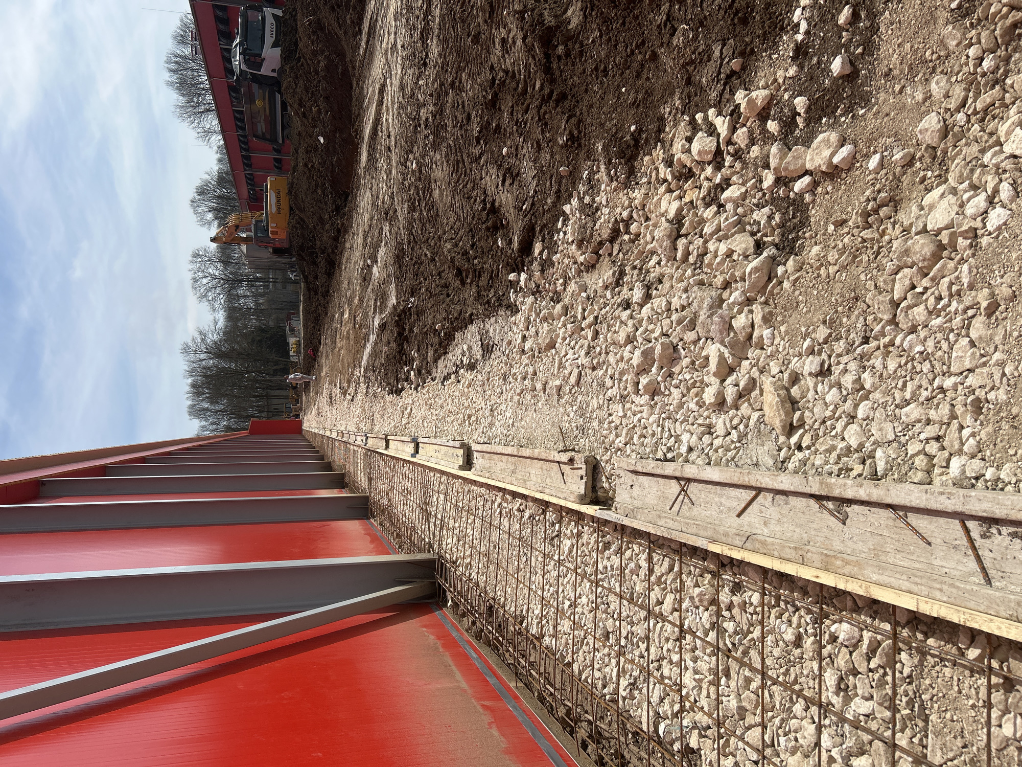 The image captures an active construction site in Donchevo, Bulgaria, under a partly cloudy sky. A large, red metal-clad building with prominent vertical grey structural support beams dominates the left side of the frame. Along the base of this structure, a prepared foundation trench contains a grid of rusted steel rebar laid over a coarse bed of light-colored gravel. Wooden formwork planks, some with exposed nails, border this rebar-and-gravel section. To the immediate right of the foundation work, the terrain transitions to dark brown, excavated soil and mud, displaying distinct vehicle tire tracks. In the mid-distance, an orange Hyundai excavator is positioned on a mound of earth, its boom raised. Adjacent to the excavator, a white Iveco dump truck is parked, facing left. A solitary individual, dressed in light-colored attire, is visible in the background near the center-left, facing away from the camera. A line of bare deciduous trees forms the distant backdrop beyond the construction zone.