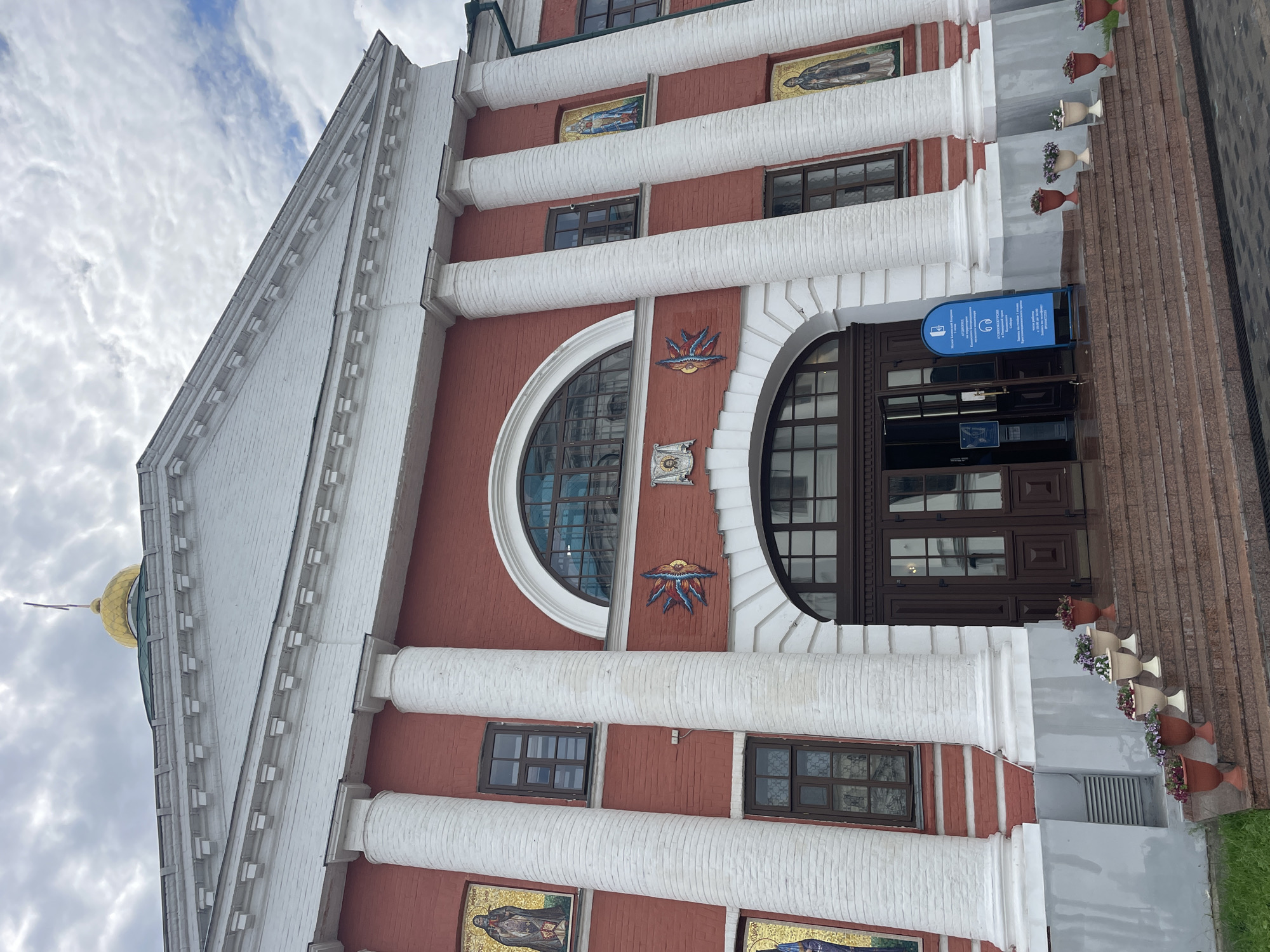 The image shows the entrance of the Kazan Kremlin's museum complex. The building is red brick with white columns and a white pediment. A golden dome is visible on the roof. The entrance features large double doors and a semicircular window above. Mosaic icons are displayed on the facade. A blue informational sign stands to the right of the entrance, and potted flowers line the steps leading up to the building. The sky is cloudy.