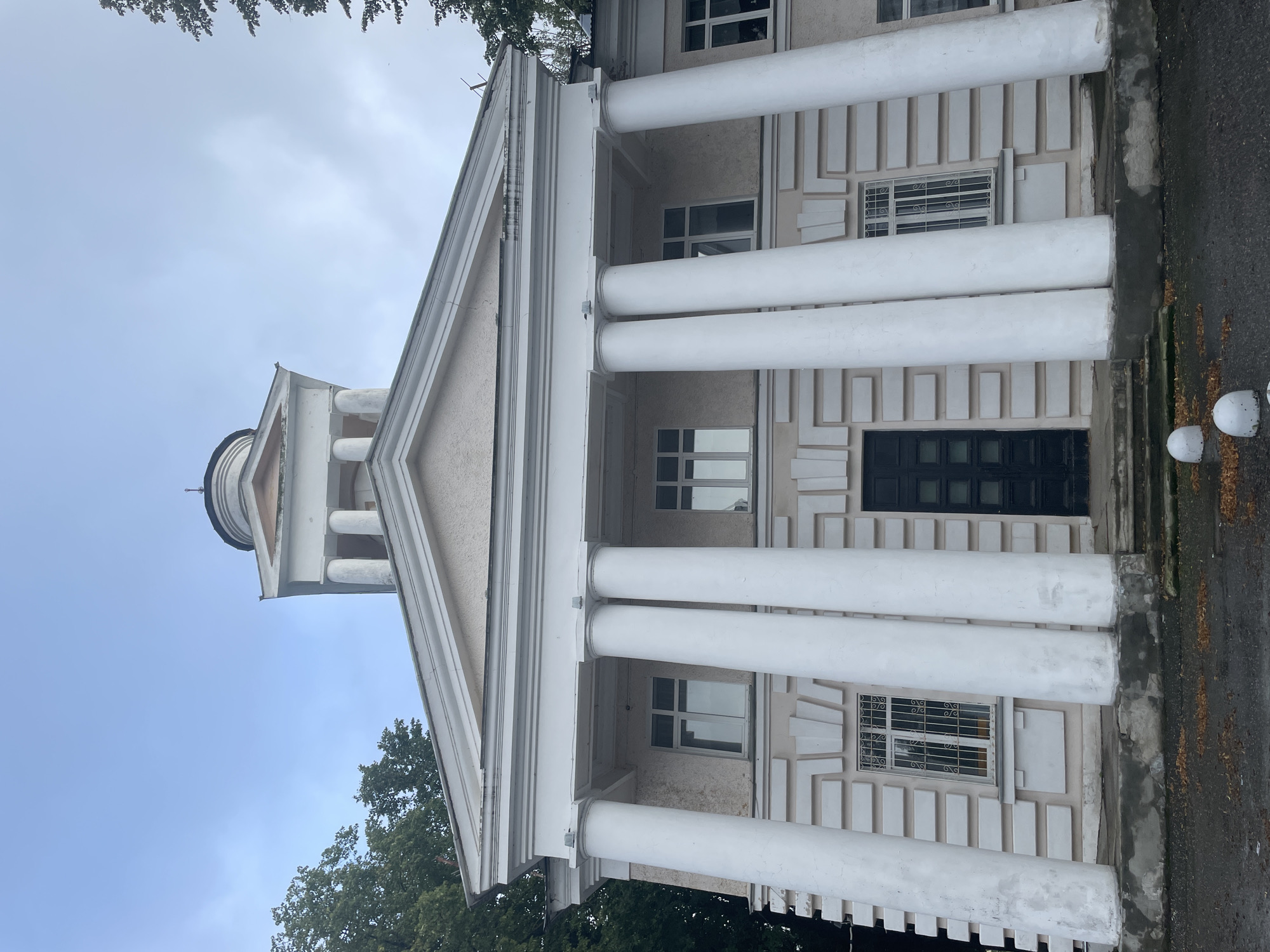 The image shows a close-up, low-angle view of the facade of a light-colored building with classical architectural elements, possibly a public building or historical site, located in Zavolzhye, Russia. The building features a prominent portico with six large white Ionic columns supporting a triangular pediment. Above the pediment, a dome-like structure topped with a spire is visible. The facade is decorated with decorative rectangular panels. There are multiple windows on the building, some with decorative metal grates. A dark double door is centered at the base of the portico. The ground in front of the building is paved and shows scattered fallen leaves. Two white spherical bollards are present in the foreground. The sky is overcast. The overall condition of the building appears somewhat weathered, with some cracks visible on the upper facade and evidence of wear on the stonework at the base. Trees with green foliage are visible on the sides of the building and partially obscure the upper structure. No people are visible in the image.