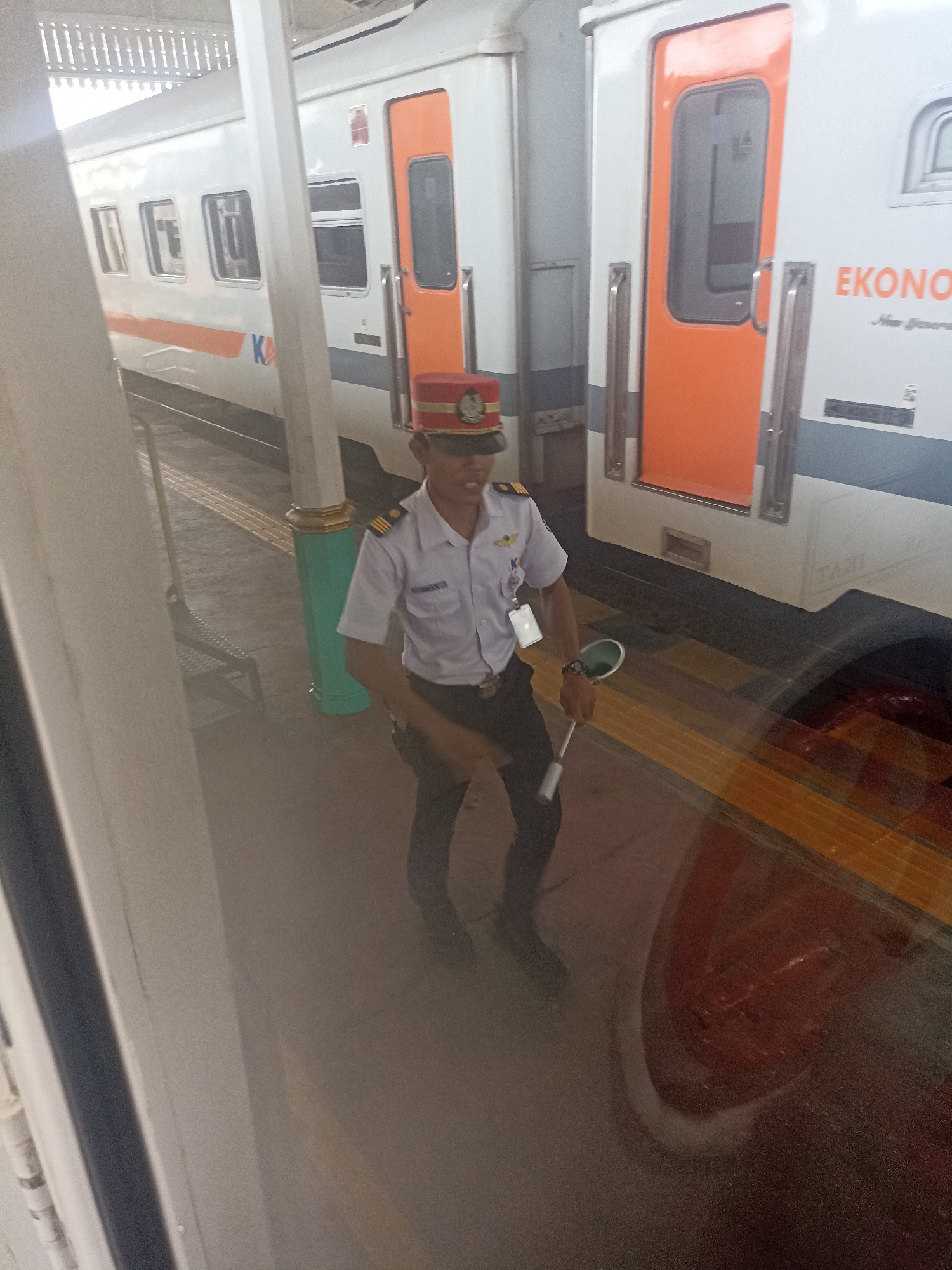 An unidentified male train conductor stands on a train platform in Probolinggo, Indonesia, holding a disc-shaped signaling device. The conductor is wearing a light blue uniform shirt, dark pants, and a red hat with a gold band. The train is white and orange, with "EKONOMI" printed on its side. The platform is paved with yellow and gray tiles. The scene appears to be a train station, with the train visible behind the conductor. The conductor is looking towards the viewer.