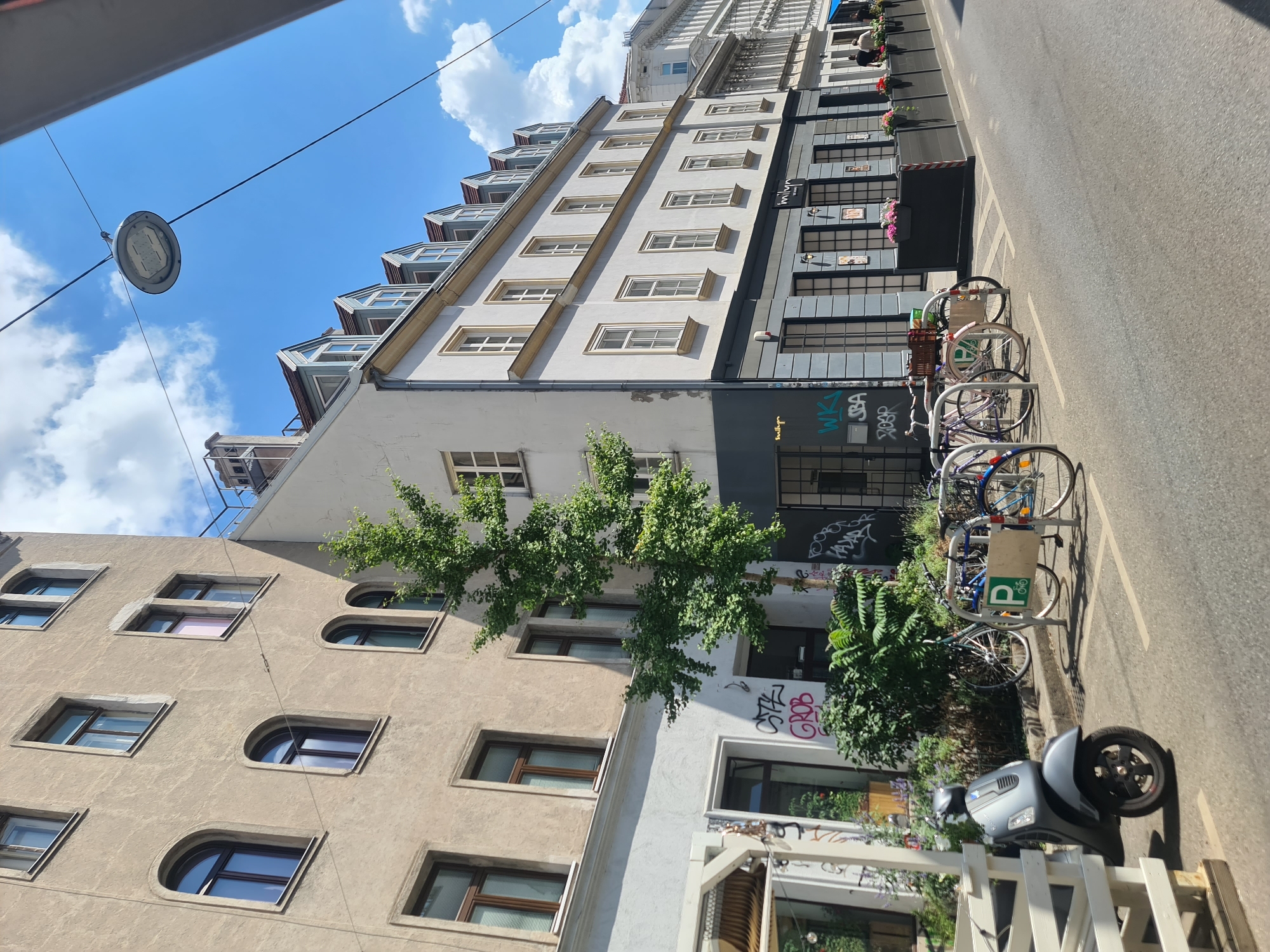 This is a street-level, eye-level, medium shot of a European city street, likely Vienna, Austria, on a sunny day. The scene features a row of buildings with traditional European architecture, including arched windows on the left building and mansard roofs on the building to the right. Several bicycles are parked in a rack in the foreground, along with a gray scooter on the left. A tree with green foliage grows between the two buildings. Graffiti is visible on the lower walls of the buildings. The street is paved and there are some flower boxes along the building on the right. Two individuals are visible walking in the distance on the right side of the frame. A street light is suspended by wires above the street.