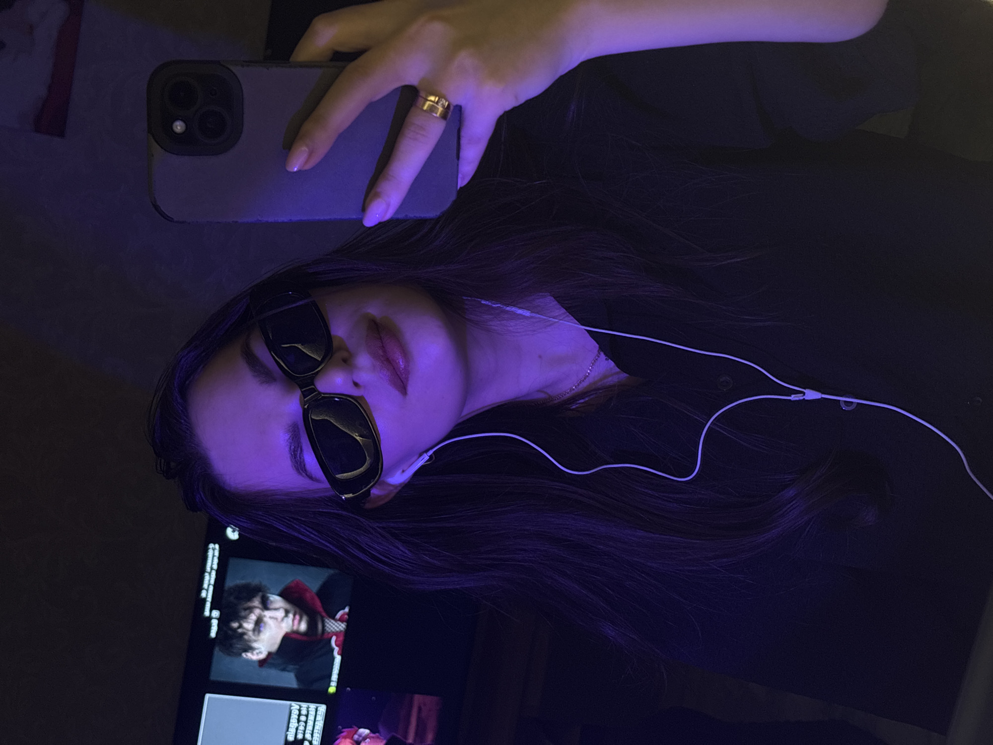 A young adult woman is taking a selfie indoors. She is wearing sunglasses and headphones and has a gold ring on her finger. She is holding a smartphone with a triple-lens camera. A screen showing a program with a male character is visible. The scene is lit with a strong purple hue. There is no information about the location of Chaikovsky, Russia.