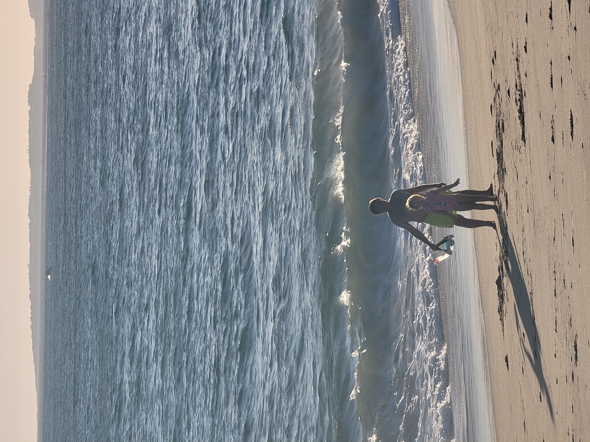 A man and a young girl stand on a sandy beach in Quarteira, Portugal. The man, wearing yellow shorts, has his arm around the girl, who is wearing a pink and white striped swimsuit. They are both looking towards the water, which is a bright blue and has small waves breaking on the shore. The sun is shining, and the light suggests it is daytime, likely late afternoon given the long shadows cast on the sand. The man is holding a snorkel and mask in his left hand. The beach appears mostly empty except for them, and the water is calm. The scene evokes a sense of peaceful family time by the sea.
