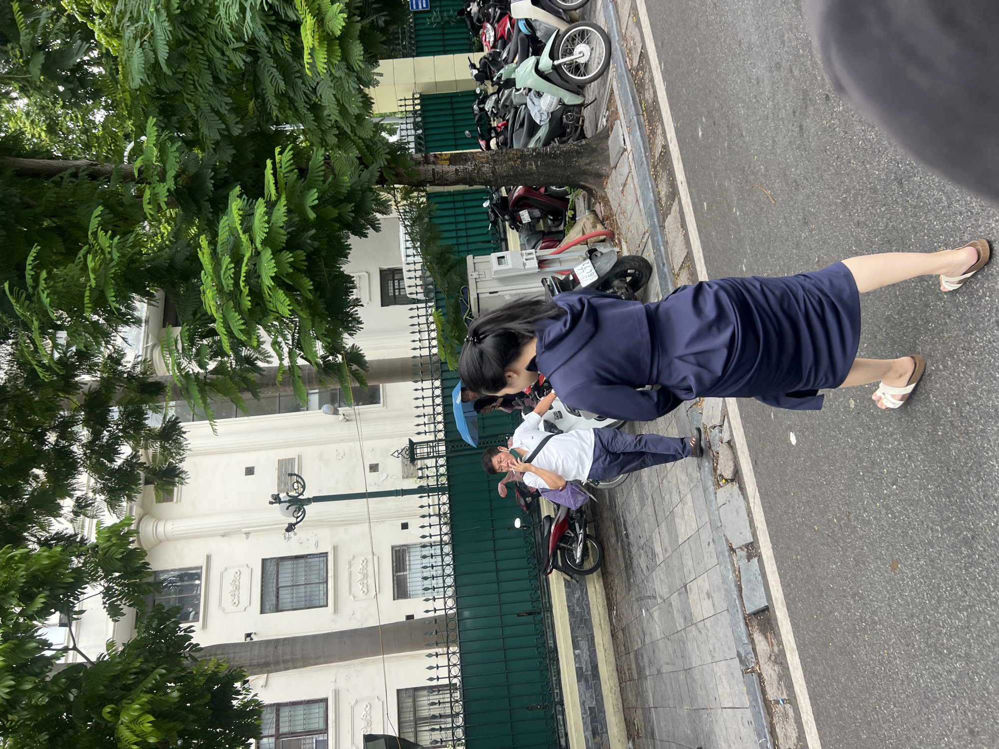 In Hà Nội, Vietnam, on a street with a sidewalk and parked motorcycles, a woman with her hair in a ponytail, wearing a dark blue hooded dress and white sandals, walks away from the camera. To her left, a man in a white polo shirt and dark pants smokes a cigarette. He holds a purple bag in his left hand and wears a crossbody bag. Behind him, an umbrella is held over two other people. The background features a building with white walls and barred windows, a green metal fence, and trees with dense green foliage. The street appears wet, suggesting recent rain.