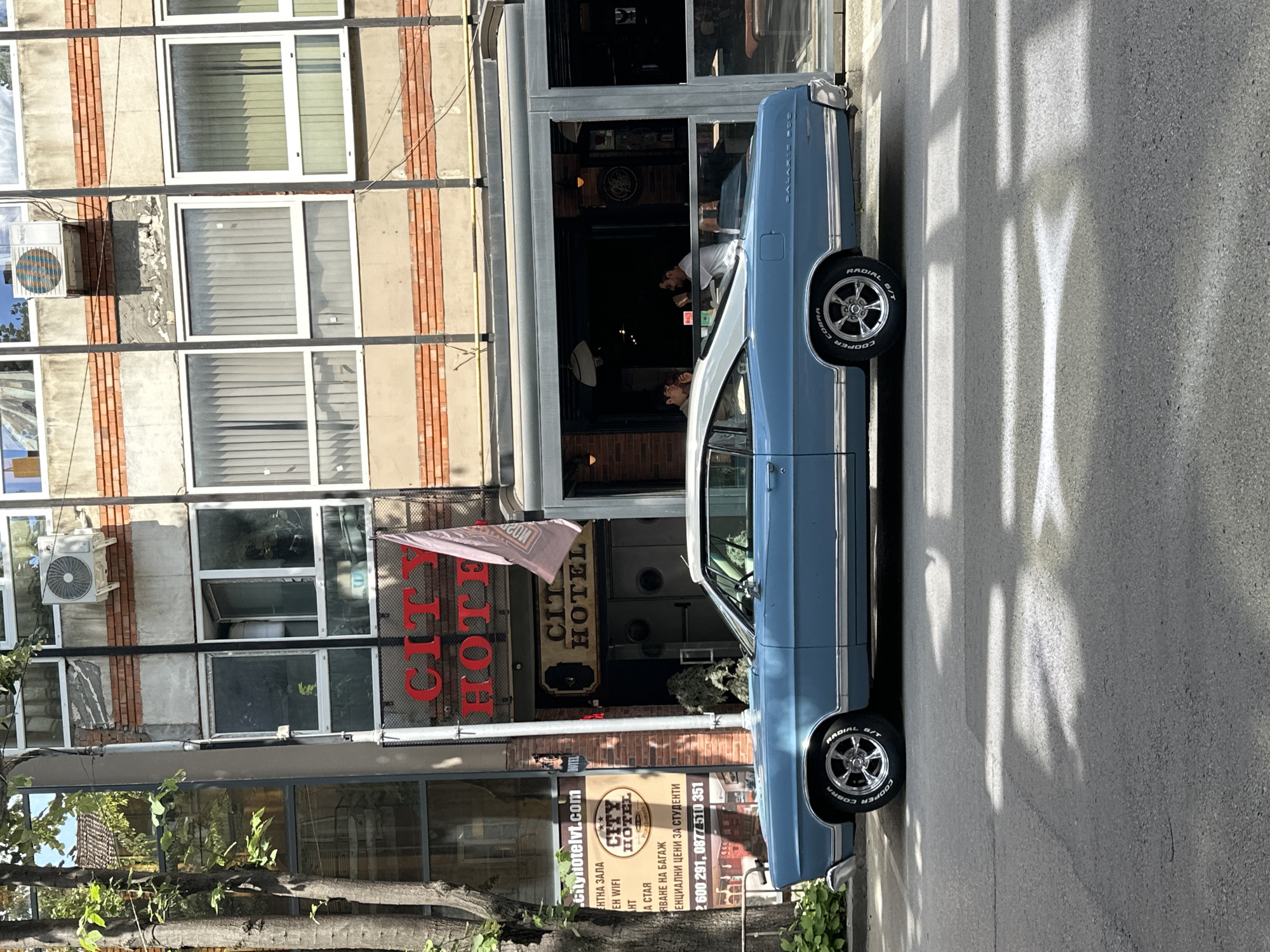 This image depicts the exterior of the "City Hotel" in Veliko Tarnovo, Bulgaria, on a sunny day. A light blue, vintage Ford Galaxie 500 is parked on the street in front of the hotel. The hotel features a brick and concrete facade with multiple windows, some of which are covered with blinds. An air conditioning unit is visible on the upper portion of the building. A flag with the hotel's name is displayed near the entrance. Inside the hotel's ground-floor establishment, visible through large glass windows, two men are seated at tables. The street is paved, and sunlight casts shadows on it, suggesting midday. Green foliage from trees is present on the left side of the frame.