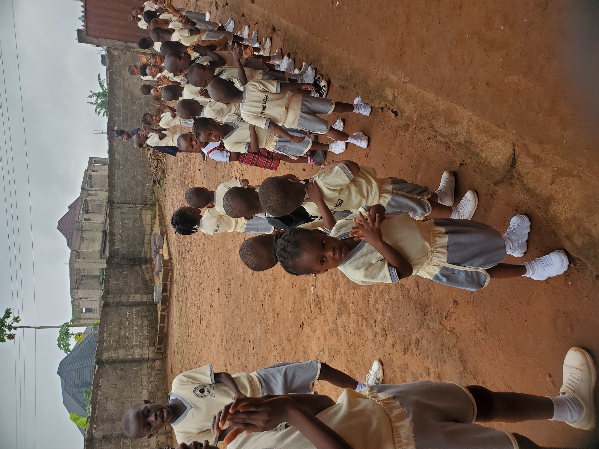 A wide, medium shot depicts a single line of children standing outside a building in Ukwu-Abwa, Nigeria. The group stretches from near the bottom of the frame towards the upper right-hand corner, comprised of roughly 30 children, presumably students. They are clad in similar uniforms, consisting of a light yellow collared shirt with dark gray sleeves and shorts, complemented by white sneakers.
The children are standing on a dirt surface that appears brown. Behind the line is a concrete wall, with a building that looks incomplete visible beyond it. A papaya tree stands close to the wall. The sky is overcast, giving the scene a somewhat muted light. The children's posture and the direction of their gaze suggest they might be addressing someone not visible in the frame.