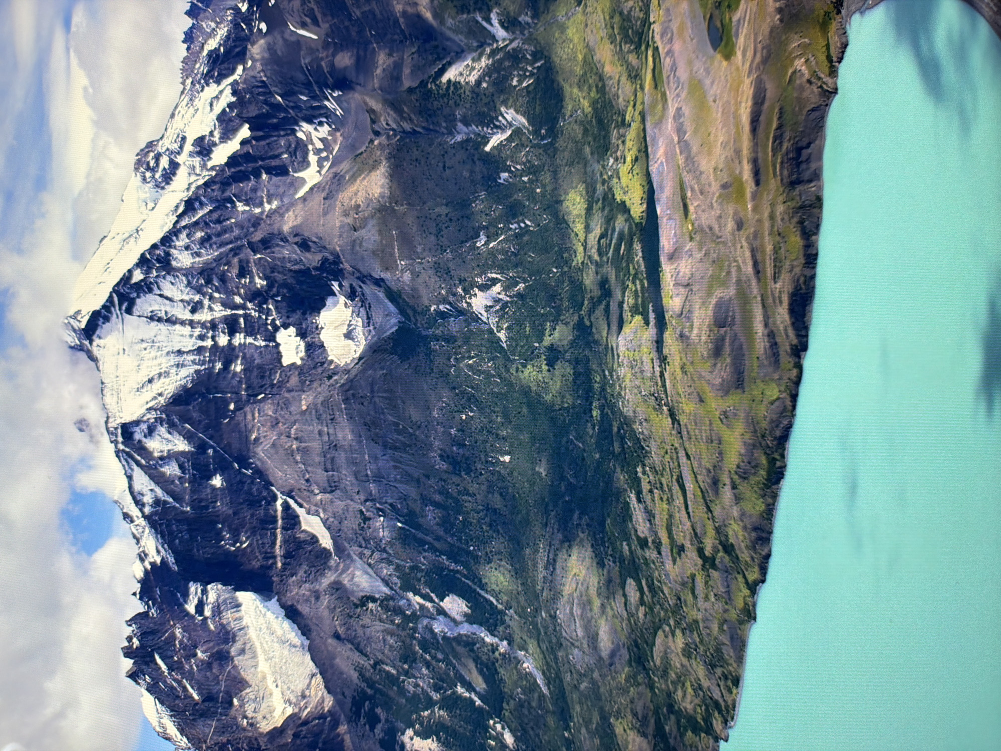 This is a landscape photograph of a vast, snow-capped mountain range towering over a vibrant turquoise lake. The mountains are rugged and imposing, with sharp peaks and deep valleys. Patches of snow cling to the upper slopes, contrasting with the dark, rocky terrain. The lower slopes of the mountains are covered in a mix of sparse vegetation and barren earth.
The lake in the foreground is a striking, almost unnatural shade of blue-green, its surface smooth and reflective. The shoreline where the land meets the water is a mix of rocky outcrops and vegetated slopes. The overall impression is one of raw, natural beauty and immense scale.
There is no information in the image to suggest any connection to the city of Kyiv, Ukraine. The scene depicts a remote, mountainous natural environment.