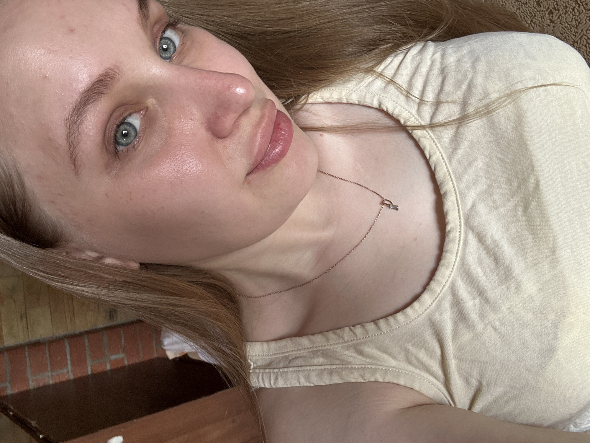 A fair-skinned, light-haired woman is the central subject of this close-up, slightly angled, selfie-style image. The woman is wearing a light-colored tank top. She has light-colored eyes and full lips. A delicate chain necklace with a small rectangular pendant adorns her neck. Her hair is parted and falls to the side, partially obscuring the background. In the lower left corner, a section of brickwork can be seen behind what appears to be a wooden surface. There is no evidence to suggest the location is Chaikovsky, Russia.