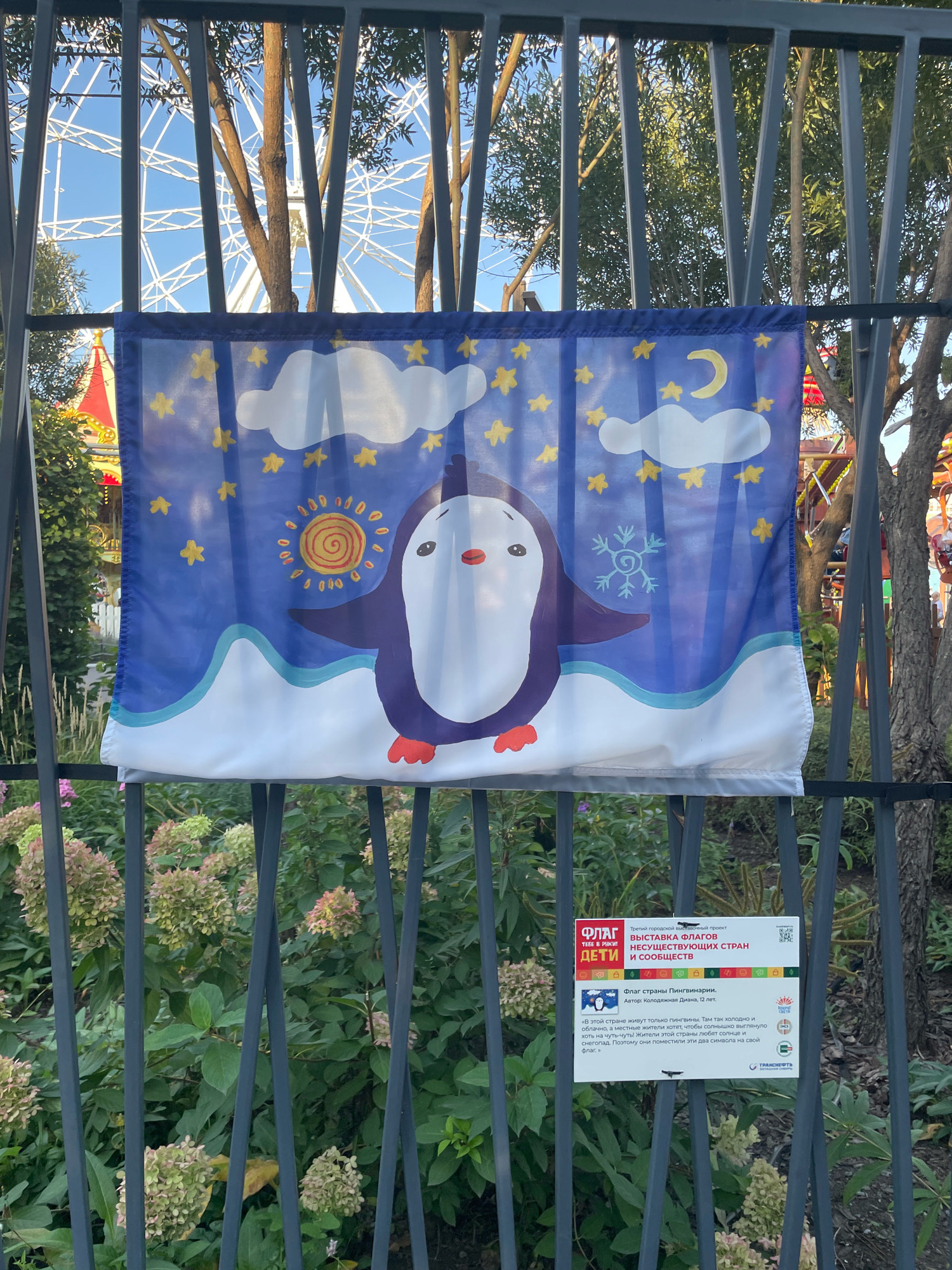An outdoor photograph shows a fence in the foreground, behind which a flag with a cartoon penguin is displayed. The flag is blue with yellow stars, white clouds, a sun, a moon, and a snowflake. The penguin is purple and white with orange feet and a red beak. To the right of the flag, a sign is attached to the fence. The sign is white and features text in Russian, as well as icons and a QR code. The sign's title reads "ВЫСТАВКА ФЛАГОВ НЕСУЩЕСТВУЮЩИХ СТРАН И СООБЩЕСТВ" (Exhibition of Flags of Non-Existent Countries and Communities). Below this, the text indicates the flag is from "Страны Пингвинарии" (Country of Penguinaria) and is authored by Kolodnaya Diana, age 12. The text further describes the country as cold, with children who love the sun and snow, and thus depicting both symbols on their flag. Behind the fence and flag, a large Ferris wheel is visible, along with trees and some foliage, including flowering plants with clusters of pink and green blooms. The overall setting appears to be a public outdoor space, potentially a park or amusement area. The information on the sign implies the location is in Omsk, Russia, as it refers to a "Third City Exhibition Project".