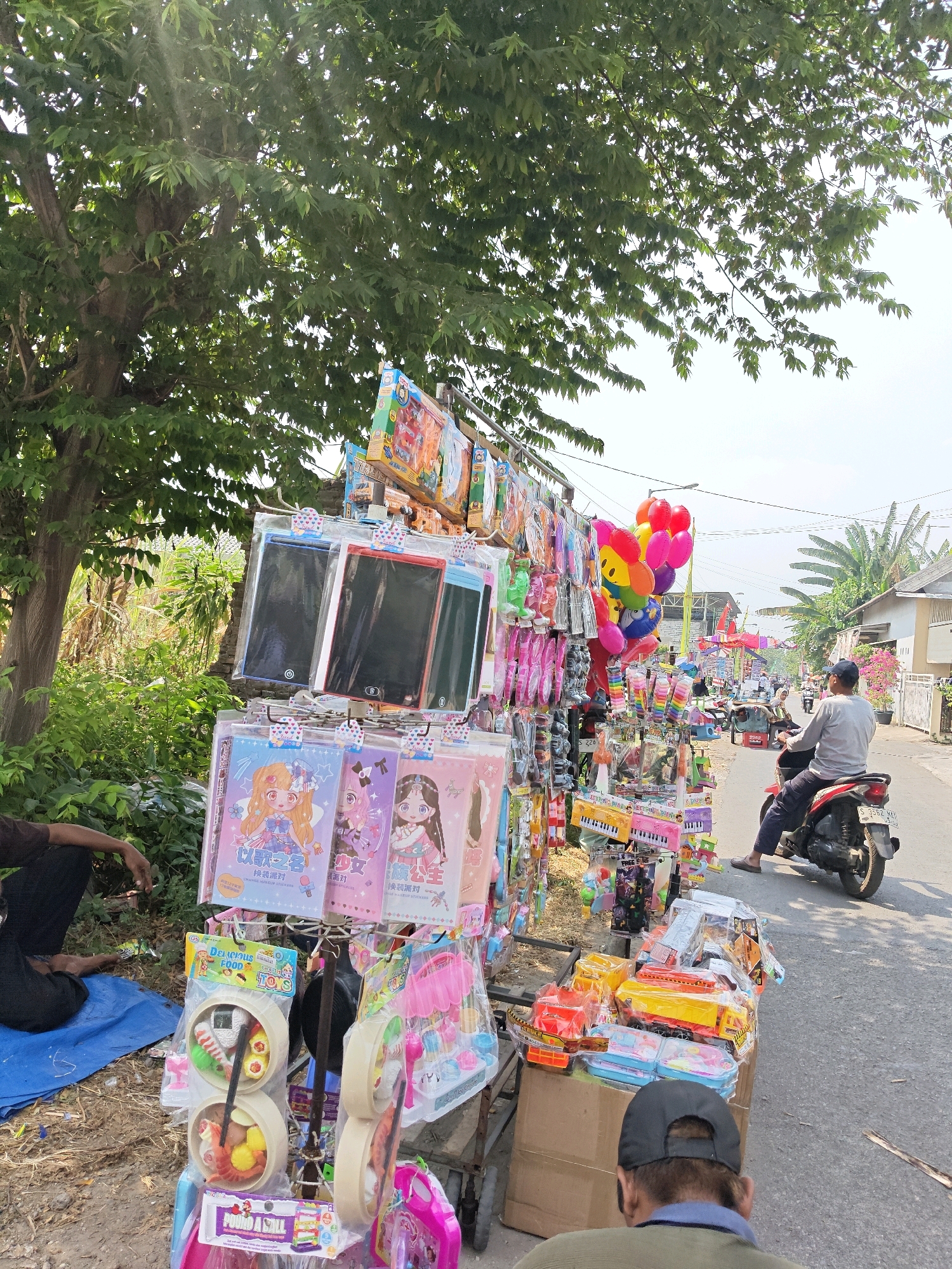 An outdoor market stall in Bangsal, Indonesia, displays a colorful assortment of toys and accessories. The stall is set up along a paved street, with a large tree providing shade over part of the scene. Several people are present, including a man sitting on a blue tarp on the left, and a person riding a red motorcycle on the right. The stall features rows of packaged toys, including what appear to be drawing tablets, stationery items, and toy food sets. Balloons in various colors, including red, yellow, and pink, are also visible. In the background, more stalls and buildings line the street, suggesting a bustling market environment. The overall impression is one of a lively street scene with a focus on children's items.