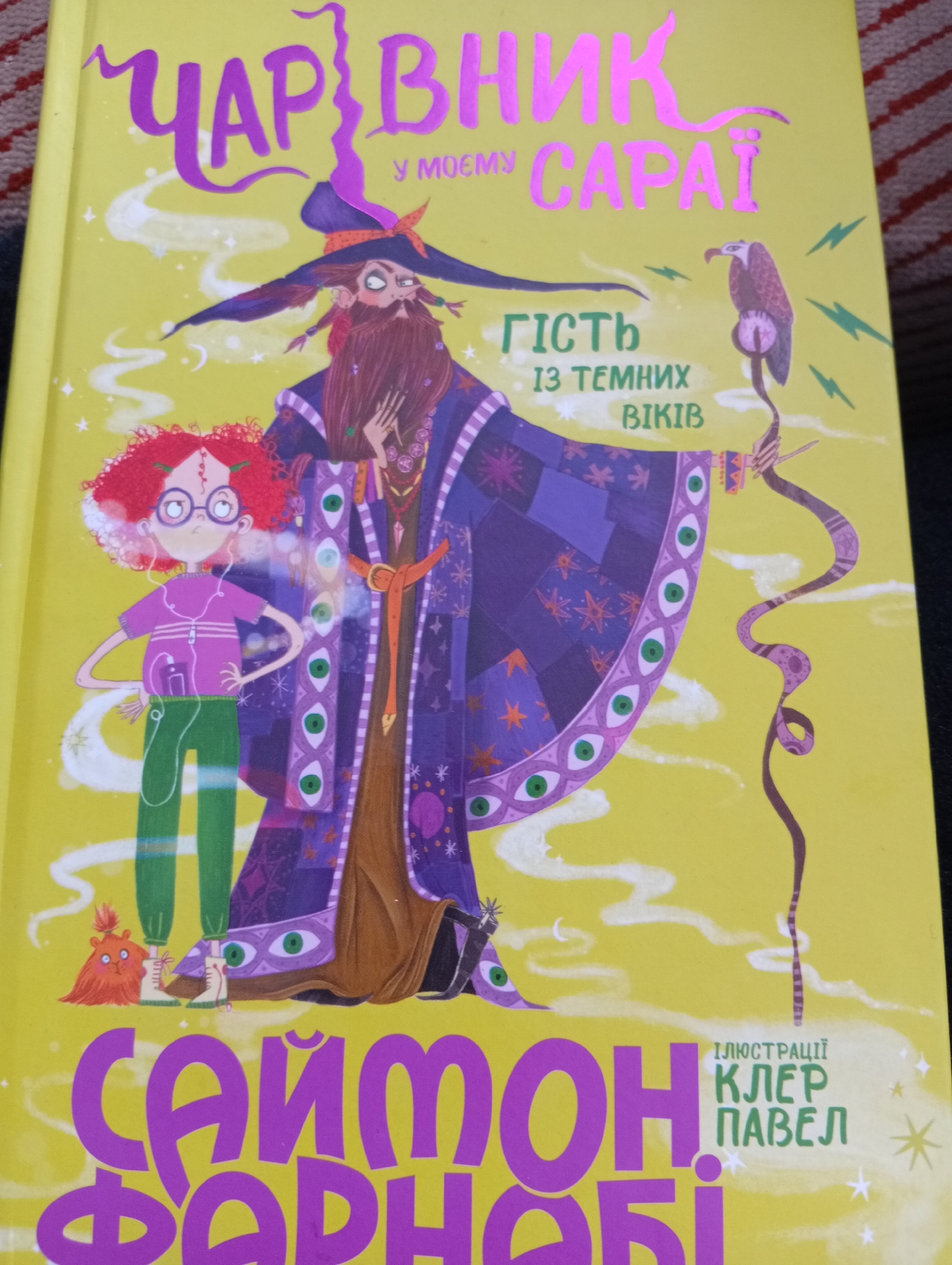 The image is a close-up, eye-level, outdoor shot of the cover of a children's book with a yellow background. The book cover features a stylized illustration of a wizard in a purple robe adorned with eye patterns and stars. The wizard has a long beard and a pointy hat. To the left of the wizard is a young girl with red curly hair, wearing glasses and headphones, and dressed in a pink shirt and green pants. A small, furry orange creature stands at the girl's feet. In the background, there are white wisps and yellow lightning bolts. The book's title, "ЧАРІВНИК У МОЄМУ САРАЇ", is written in large, purple, stylized letters at the top. Below the wizard's image, smaller text reads "ГІСТЬ ІЗ ТЕМНИХ ВІКІВ" and "ІЛЮСТРАЦІЇ КЛЕР ПАВЕЛ". The bottom of the book cover shows a large, purple, stylized title "САЙМОН ФАРНАБІ". The book is likely part of a series.