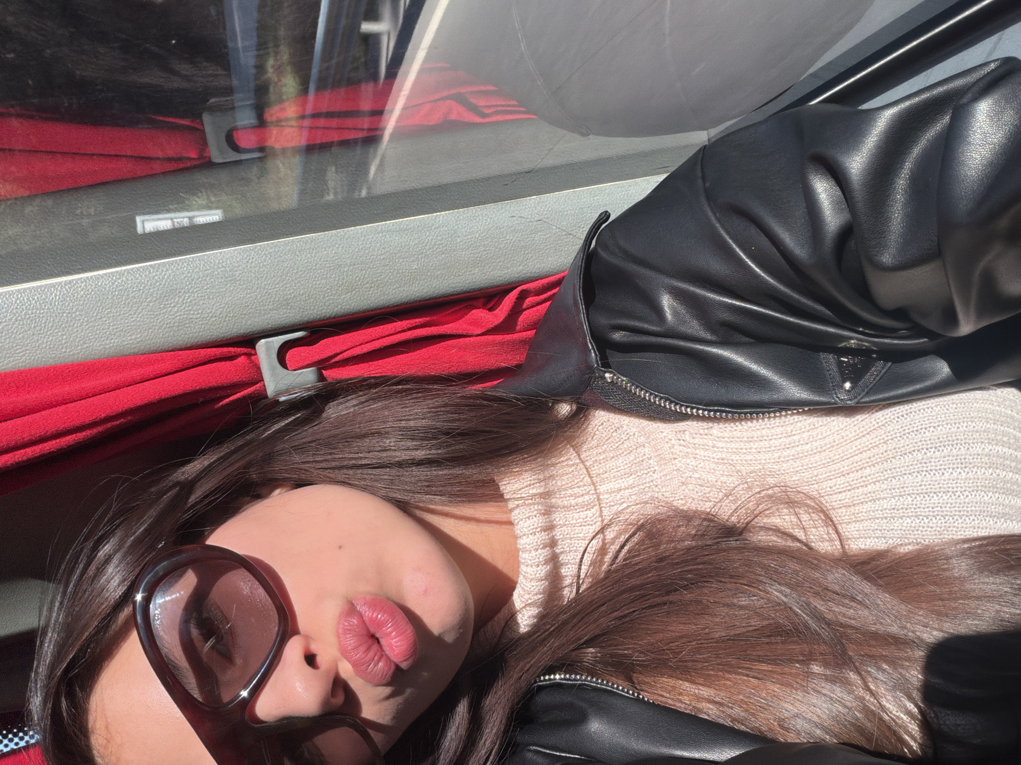 A woman with long brown hair is lying down indoors, possibly in a vehicle. She is wearing sunglasses, a light-colored sweater, and a black leather jacket. A red curtain is visible behind her head. The location may be Dolno Osenovo, Bulgaria.