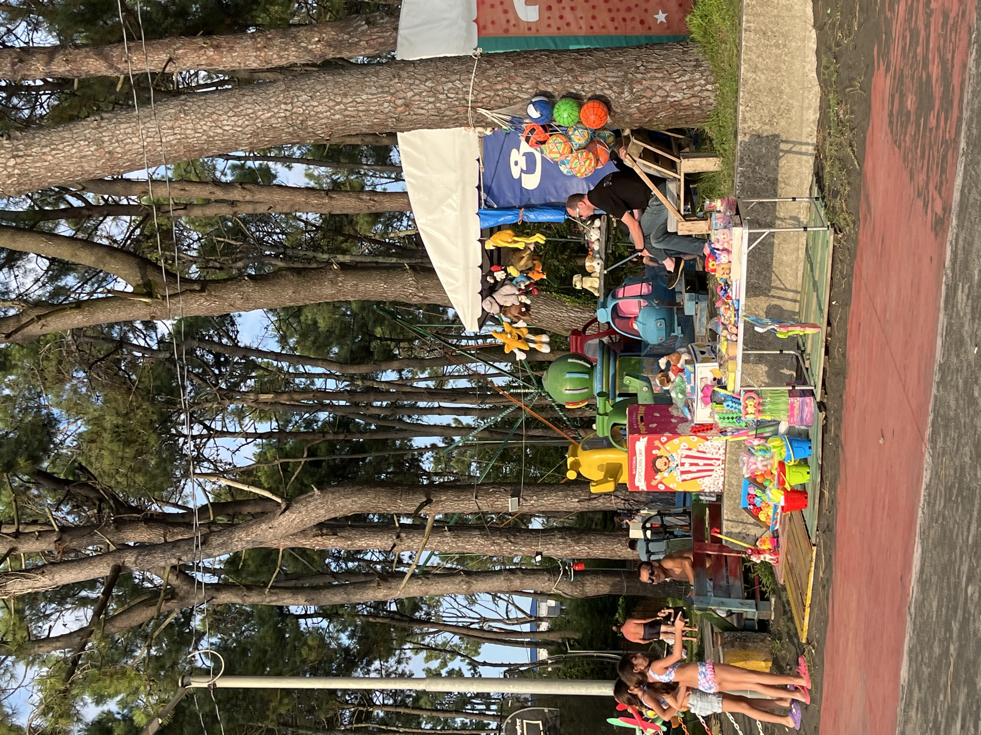 The image depicts a street scene in Ureki, Georgia, a resort town known for its magnetic black sand beaches. The foreground features a red surface, possibly a sports court or walkway, with a sandy area to the left. A row of tall pine trees dominates the background, casting long shadows and creating a natural canopy.
In the mid-ground, a collection of amusement attractions and vendor stalls are set up. To the far left, three young girls in swimwear are standing near a small playground structure. To their right, a man in sunglasses is seated on a bench. Further to the right, a white tent serves as a canopy for a vendor's stall, displaying an assortment of plush toys hanging from a line. A man in a black t-shirt and blue jeans is seated on a wooden chair, facing away from the camera, at the stall. In front of the stall, a white folding table is laden with various toys, including buckets, shovels, and small inflatable items.
Several amusement rides are visible, including a bright yellow helicopter-shaped ride and a green circular ride. A large, brightly colored box with "NATIONAL POPCORN DAY" printed on it is prominently displayed near the vendor's stall. The ground is a mix of sand and dark soil, with some wooden planks laid out as pathways. The overall atmosphere suggests a lively recreational area, possibly a boardwalk or a park near the beach. The lighting indicates it is late afternoon or early evening, with the sun casting a warm glow through the trees.